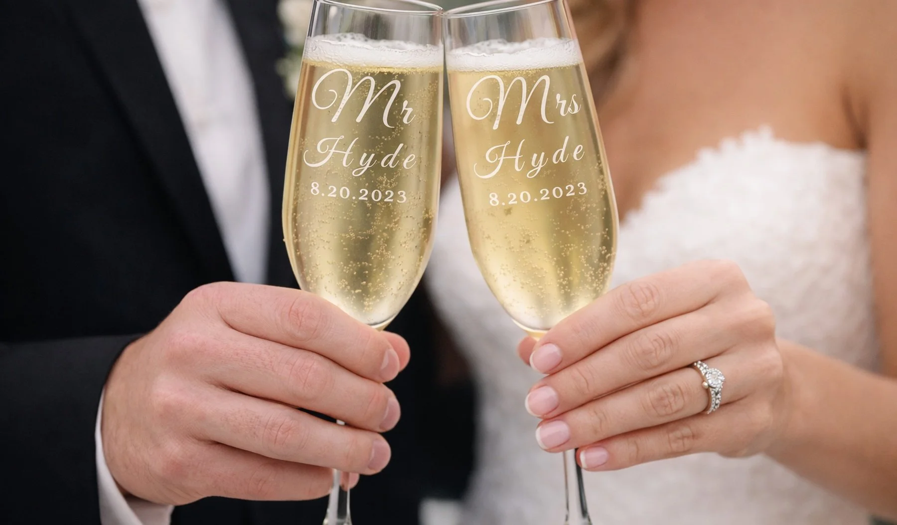 Two people dressed in wedding attire toasting with champagne flutes that have wedding details engraved on them: 'Mr. Hyde 8.20.2023' and 'Mrs. Hyde 8.20.2023'.
