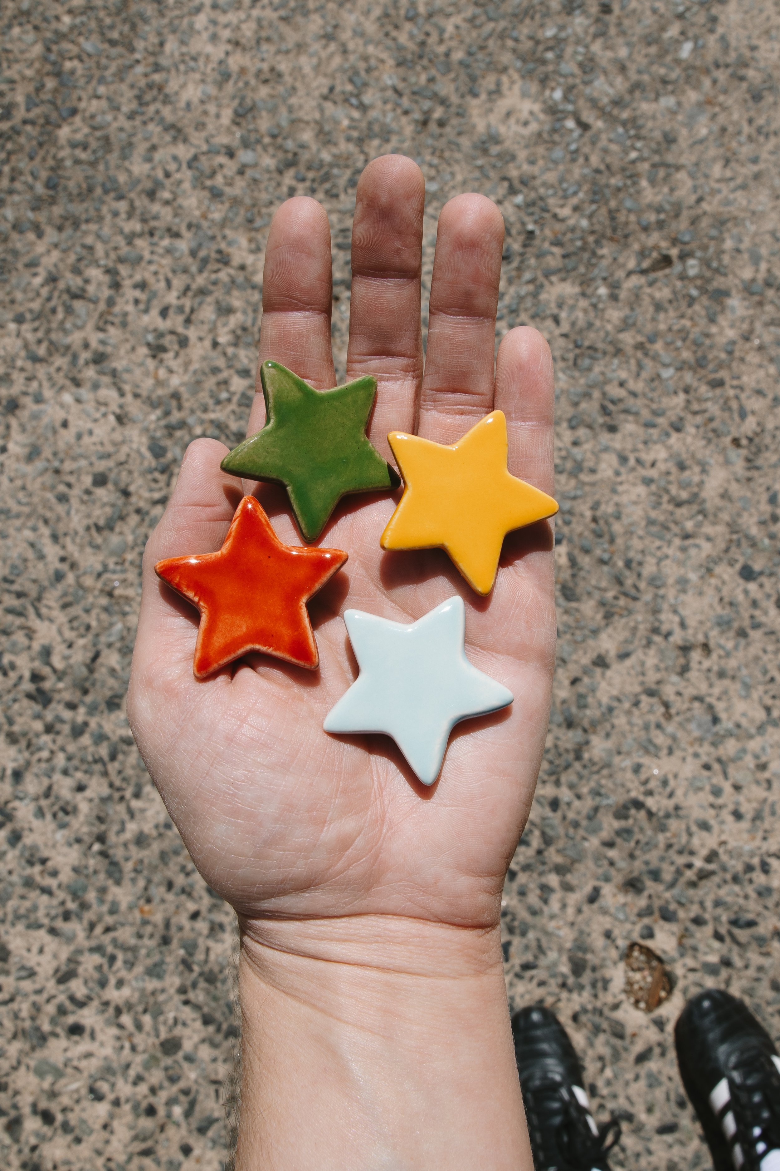 Hand holding four star-shaped ceramic objects in green, yellow, orange, and white colors, outdoors on a textured ground, with black shoes visible at the bottom of the image.