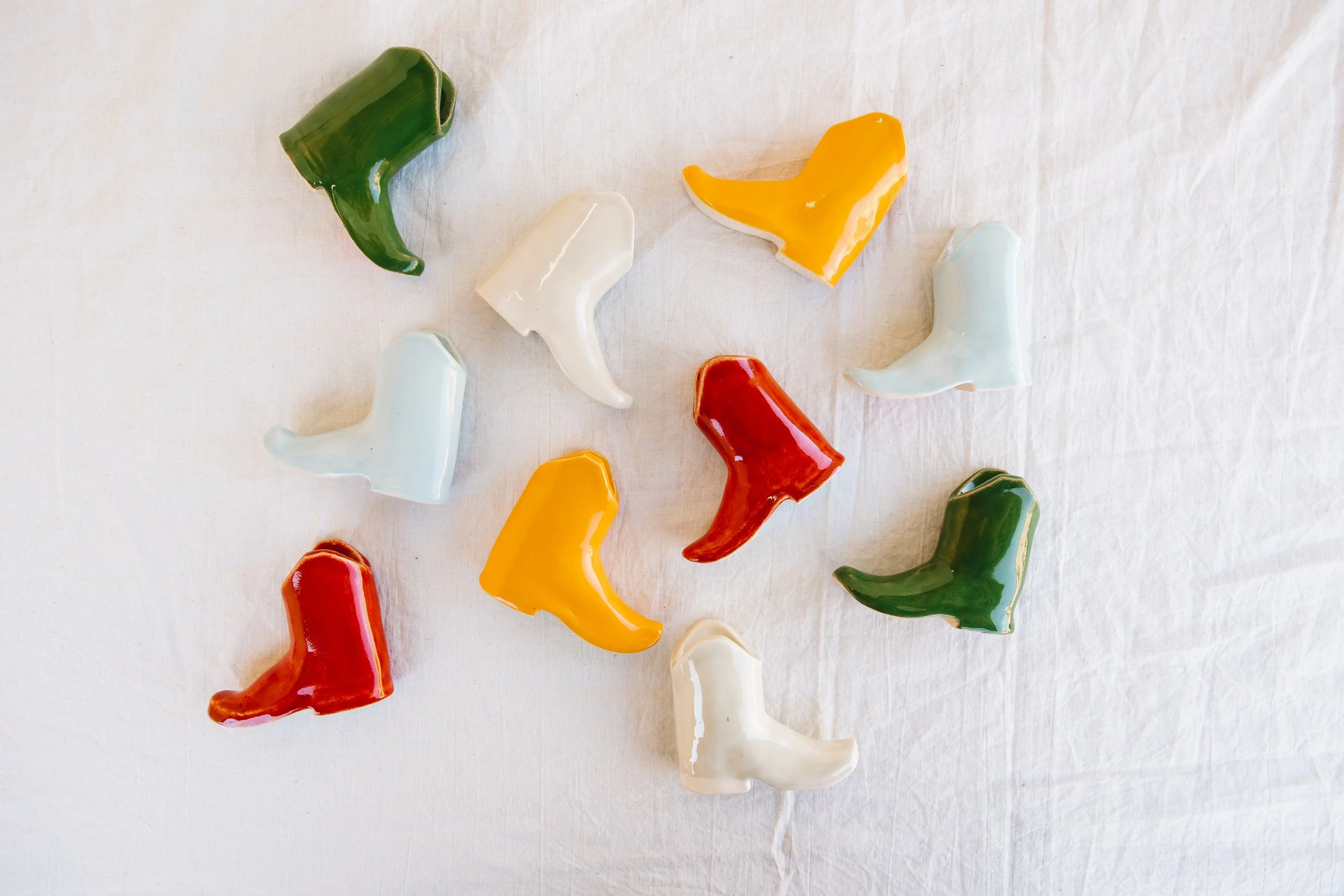 Colorful ceramic cowboy boot figurines in green, white, yellow, and red arranged on a white fabric surface.