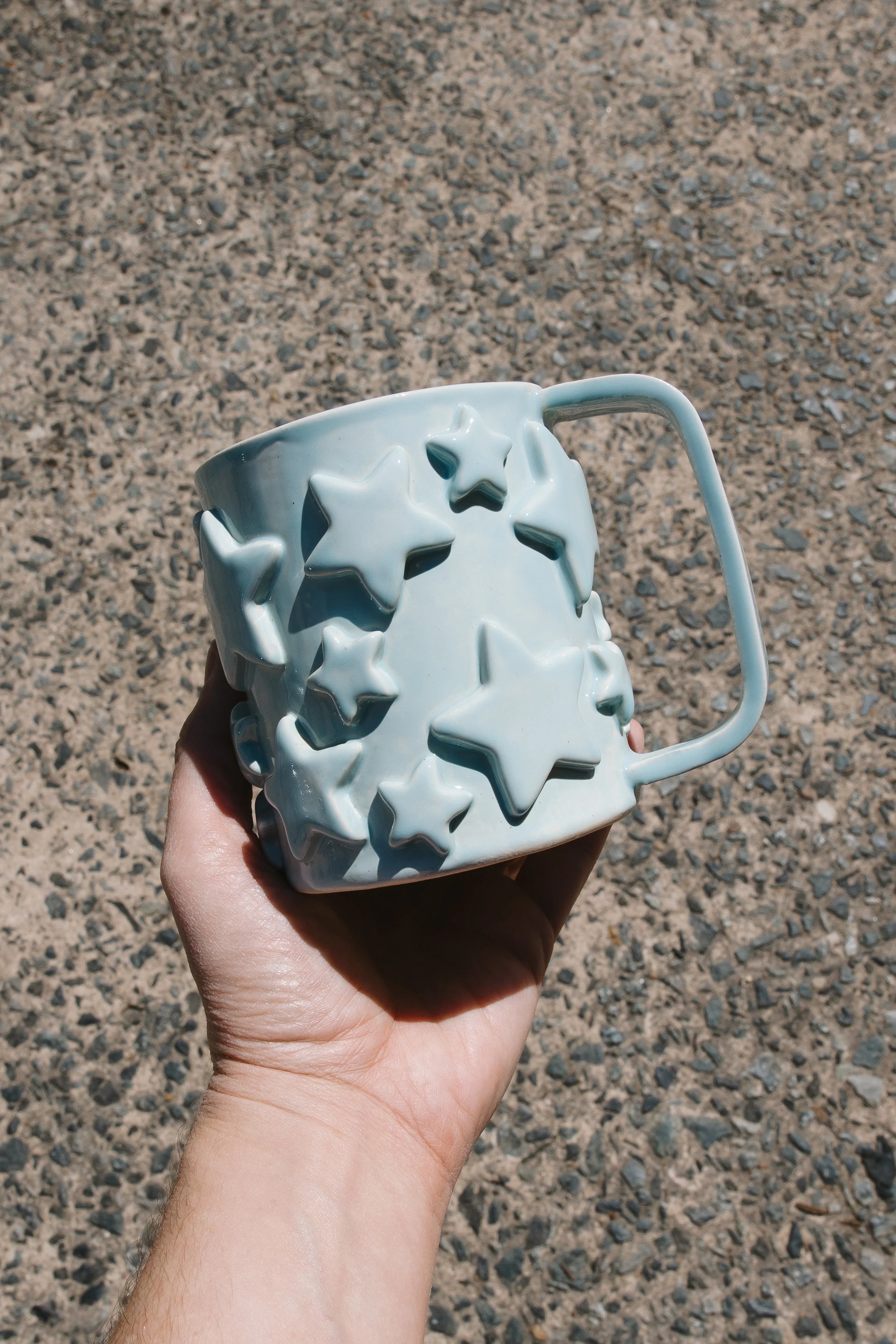 A hand holding a white mug decorated with raised star shapes, against a rough, rocky ground.