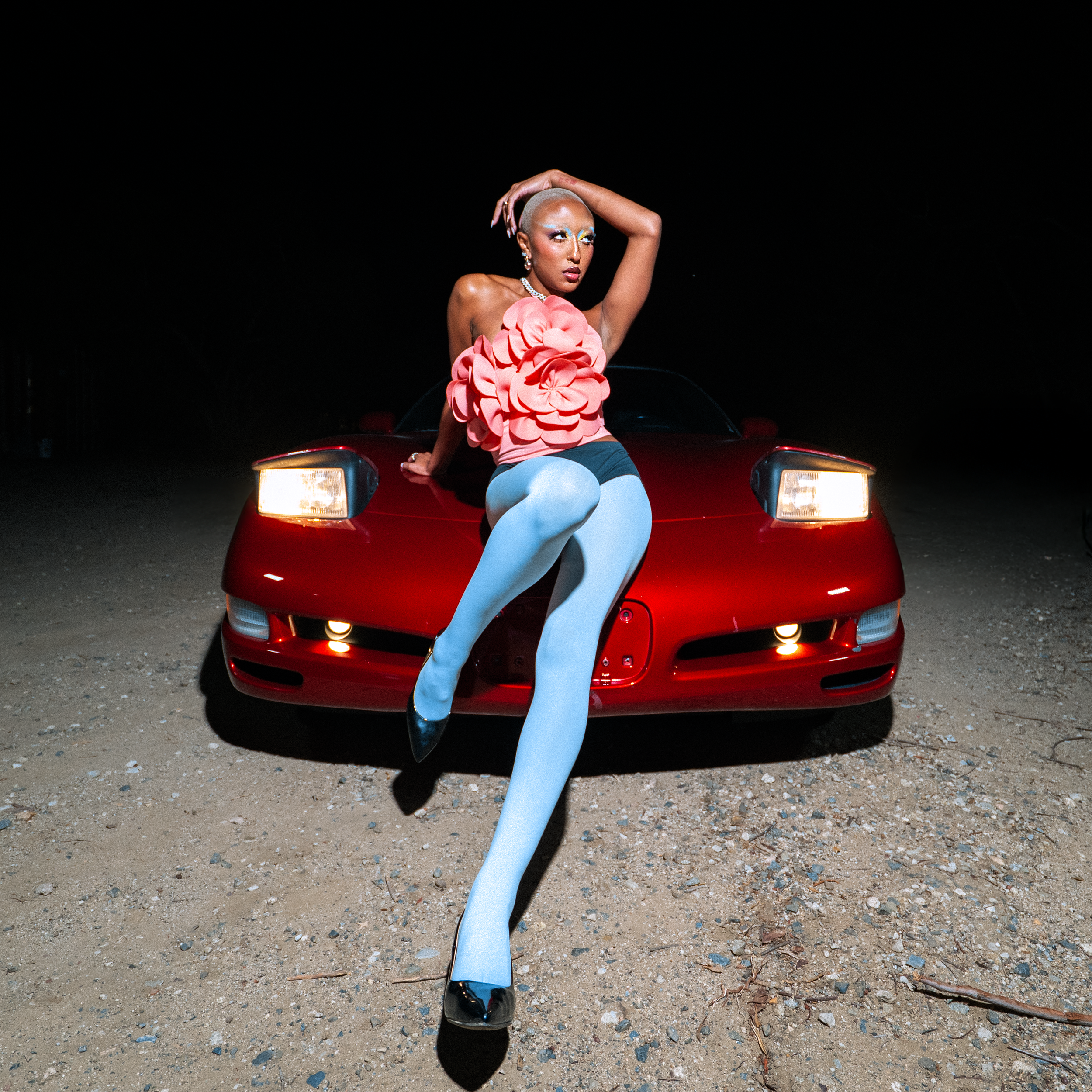 Person with makeup and platinum blonde hair posing on the front of a red sports car at night, wearing a pink ruffled top, black shorts, white tights, and black heels.