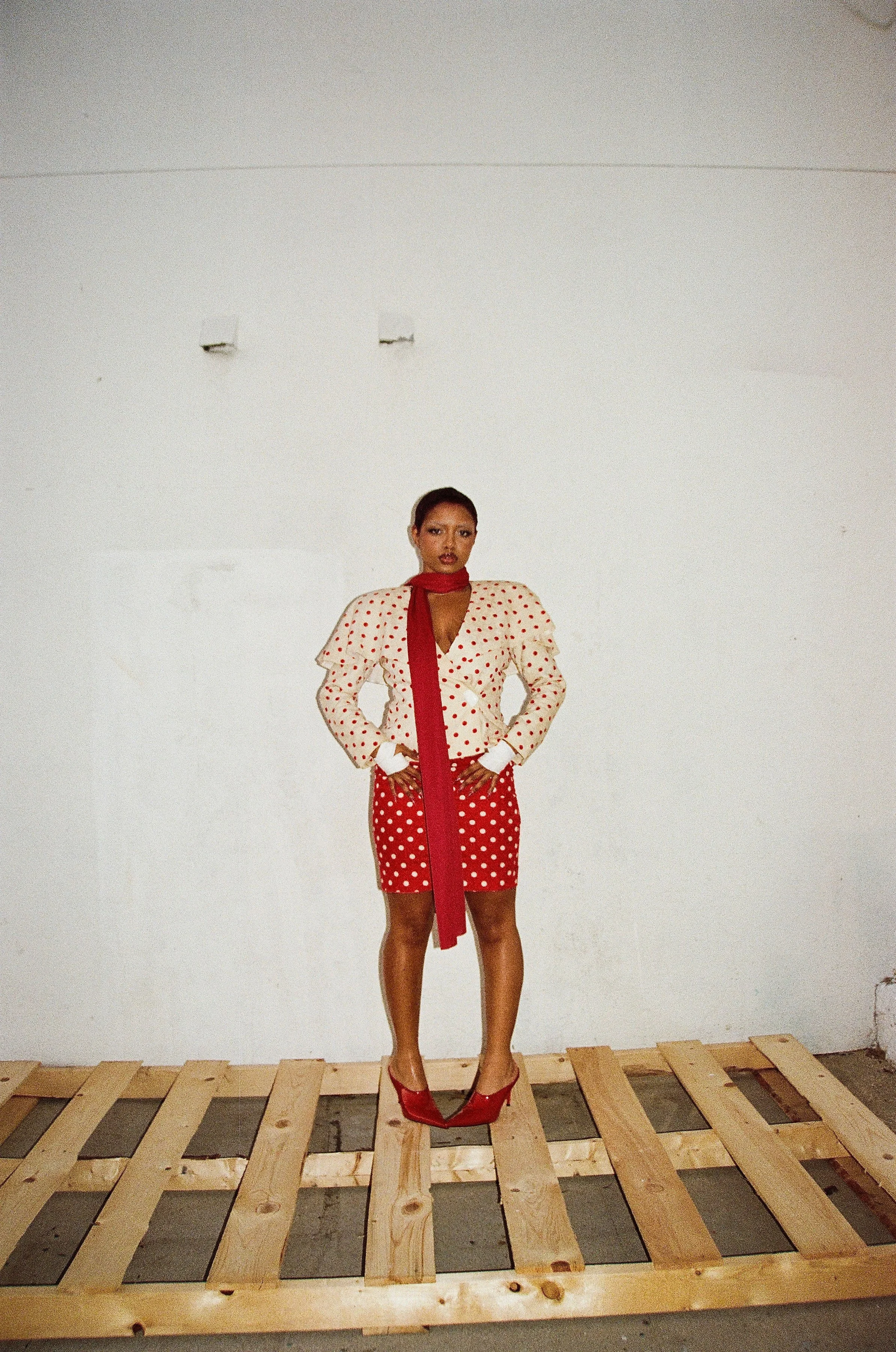 A woman standing on a wooden pallet floor against a plain white wall, wearing a polka dot dress with a matching polka dot skirt, red high heels, and a red scarf.