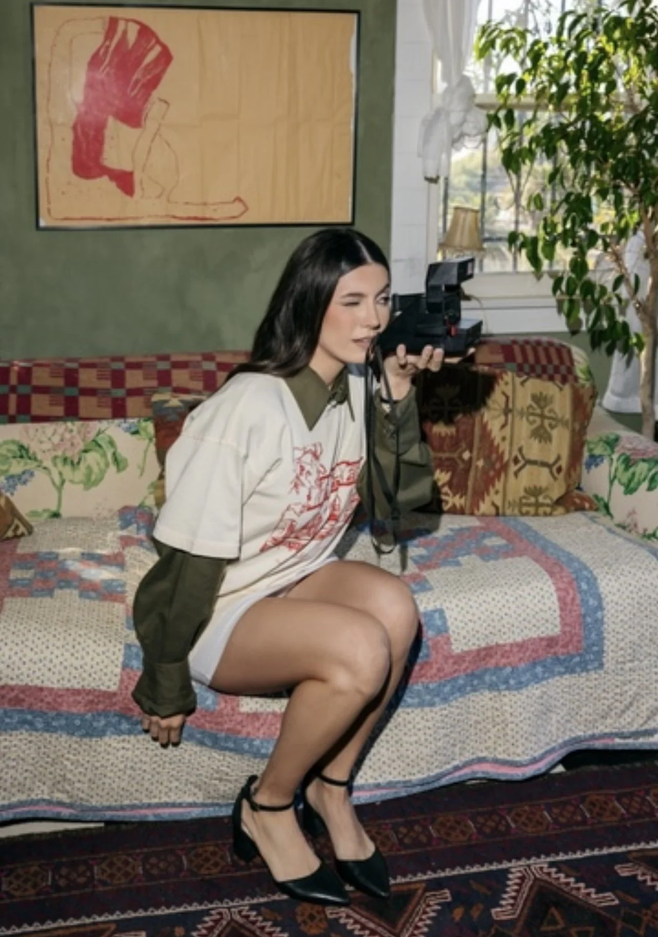 A woman with dark hair and light skin, sitting on a patterned couch, holding a vintage camera to her eye, one eye closed, in a cozy room with natural light, green walls, artwork, a bookcase, and a large window with plants.