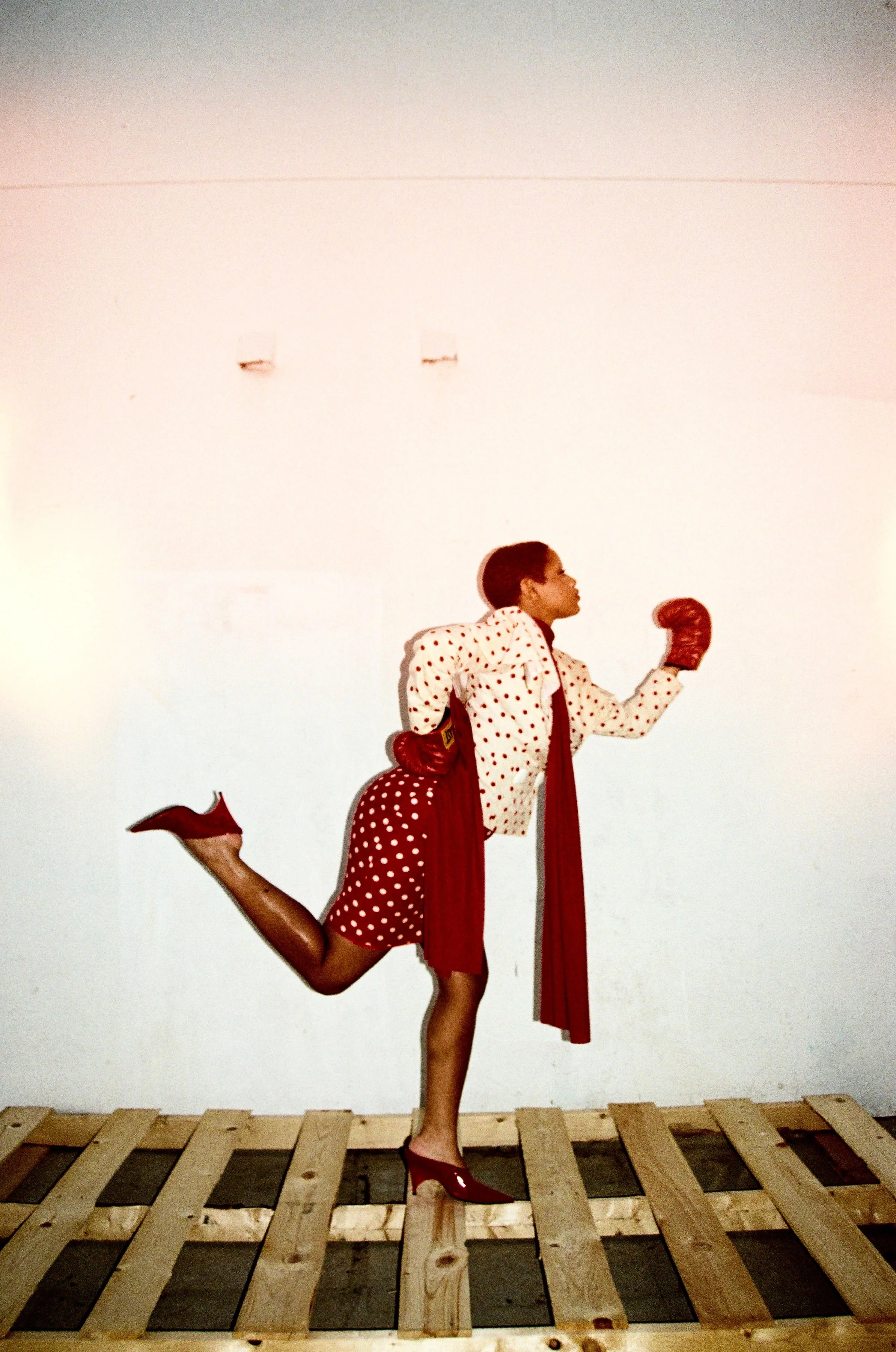 A woman with short hair dressed in vintage polka dot clothing and red shoes, posing as if running on a wooden pallet floor against a plain wall.