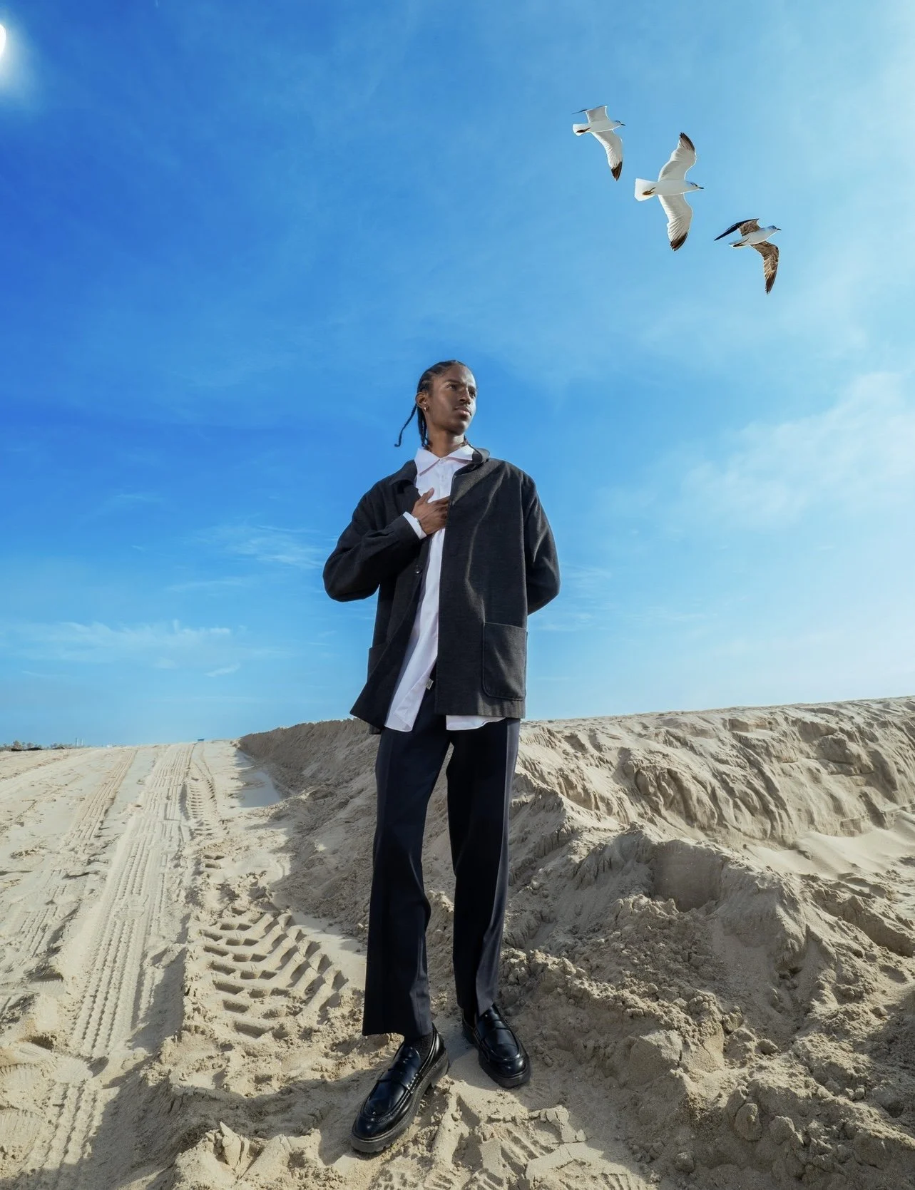 A woman standing on sandy terrain with a clear blue sky above her, with three seagulls flying overhead.