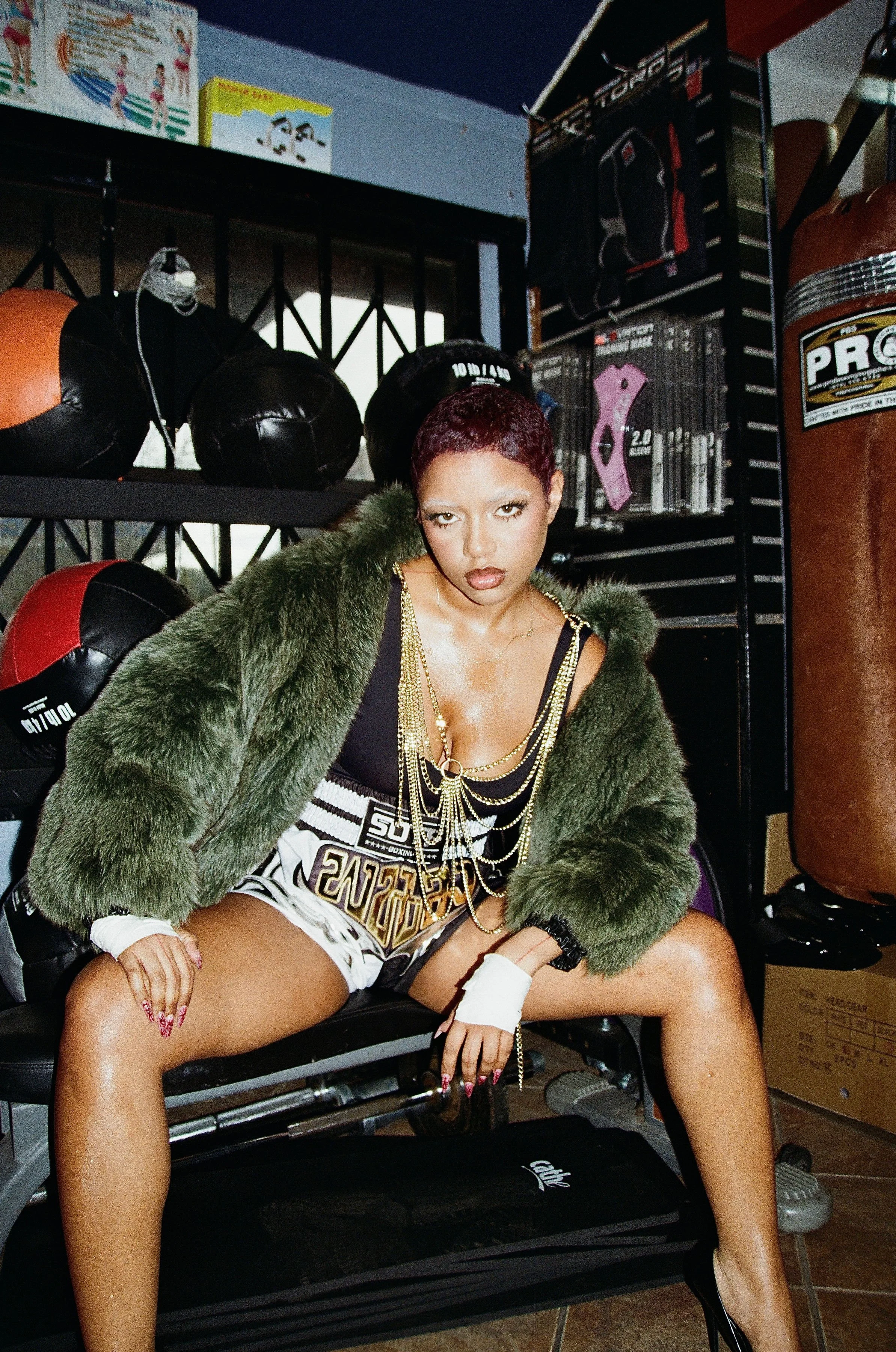 Young woman with short hair and makeup sitting on a bench in a gym, surrounded by punching bags and boxing equipment, wearing a black top, shorts, a green fur jacket, and multiple gold chains.