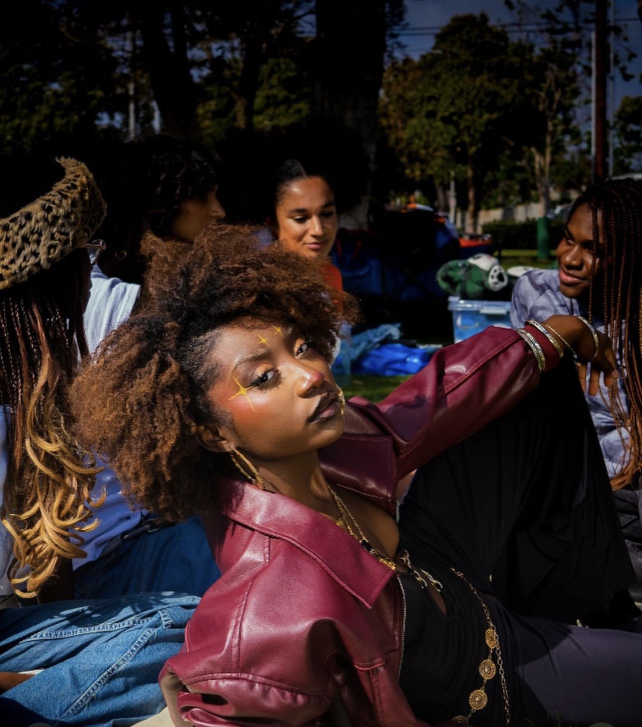 A group of women sitting outdoors during what appears to be evening, with one woman with natural curly hair and star-shaped face paint looking at the camera.