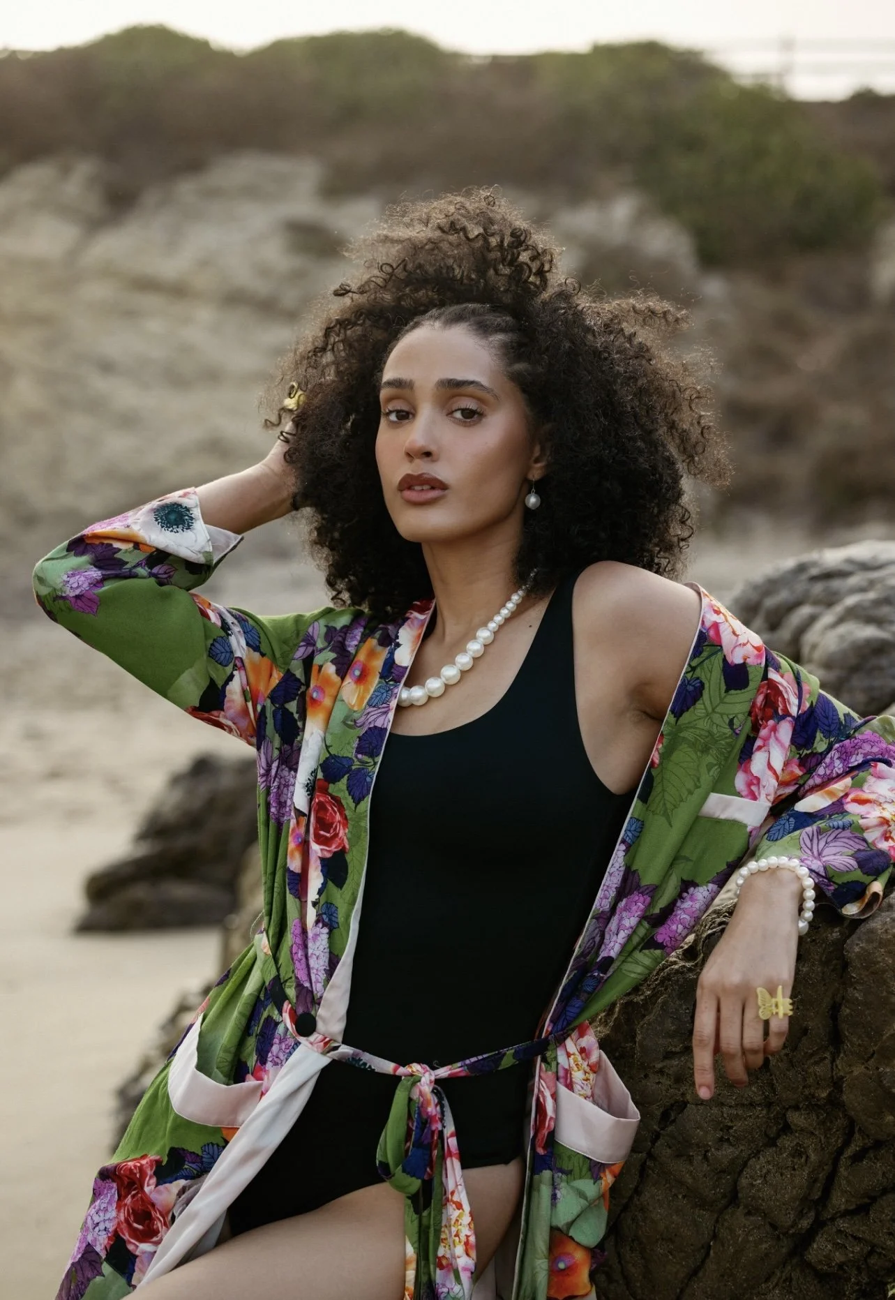 A woman with curly hair and pearl jewelry standing outdoors on rocks, wearing a black tank top and a colorful floral kimono.
