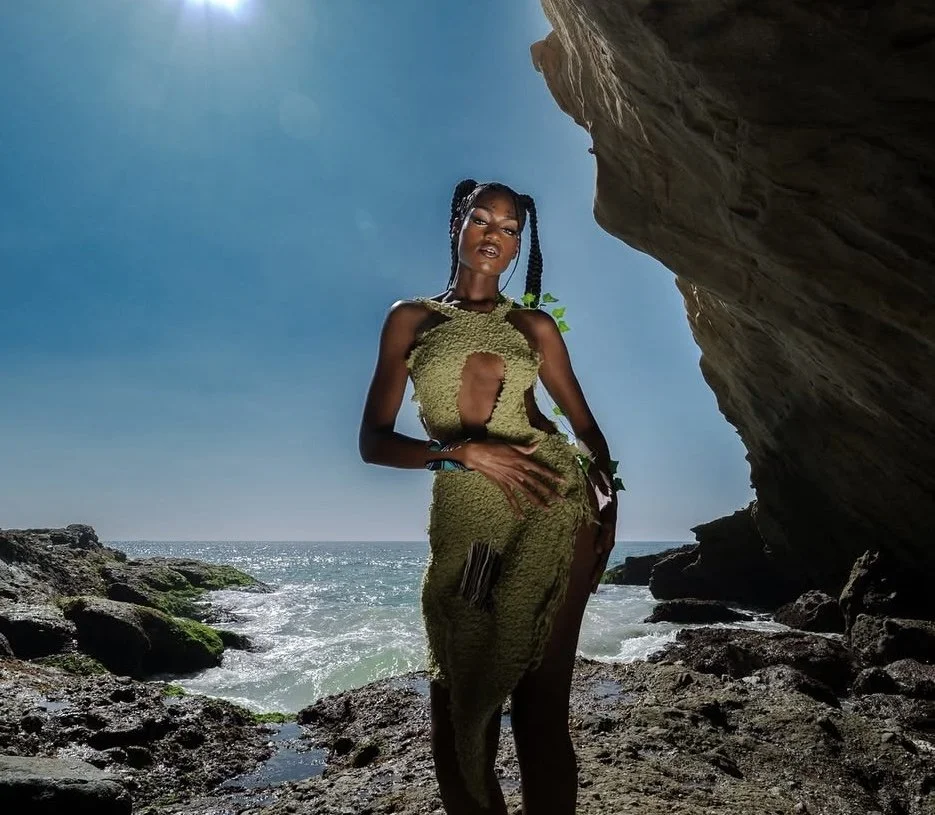 A woman in a green dress posing on a rocky beach with the ocean and a clear blue sky in the background.