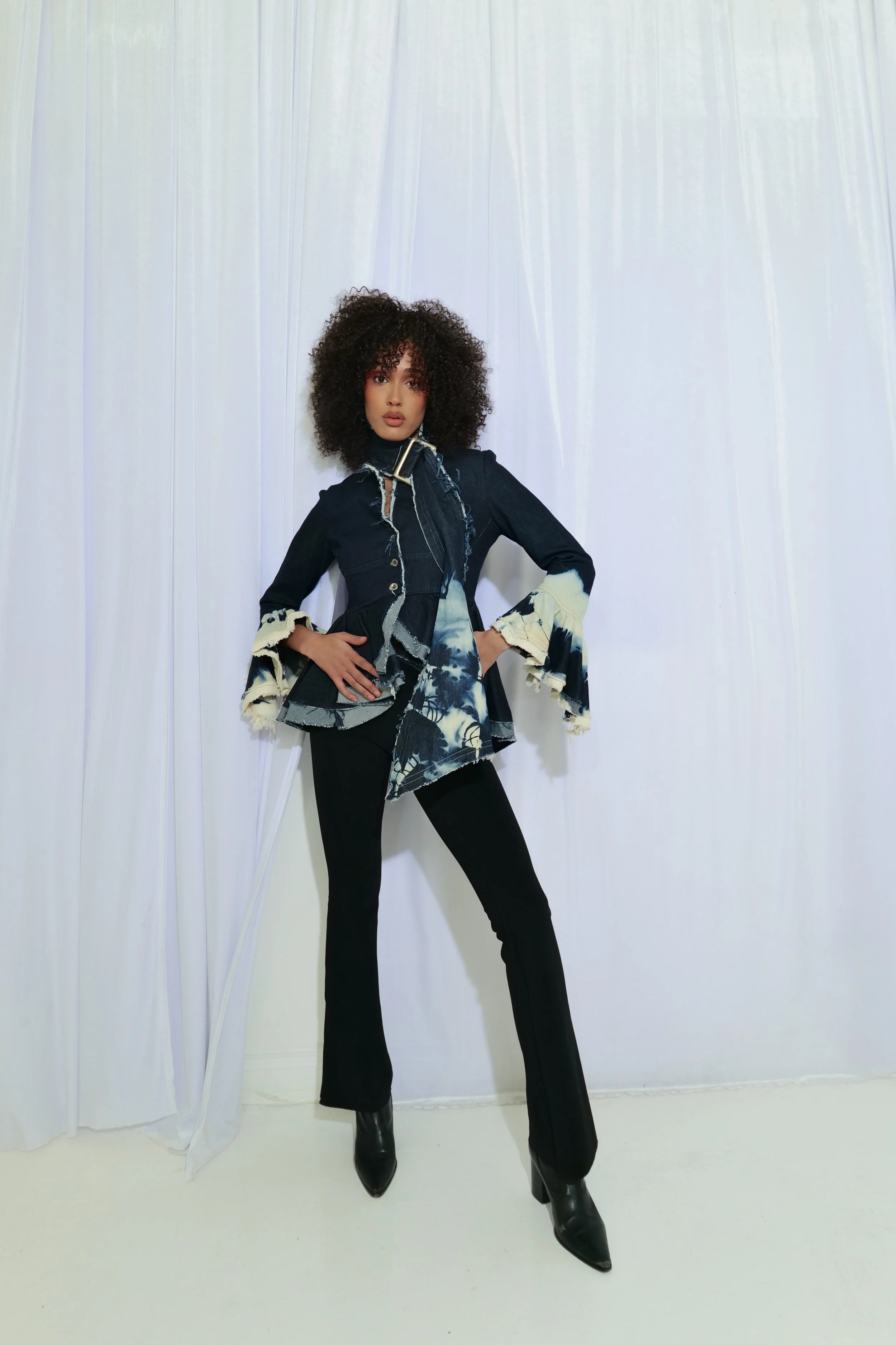 A woman with curly hair wearing a dark denim jacket with frayed bell sleeves, a patterned asymmetrical top, black pants, and black high-heeled boots standing against white curtains.