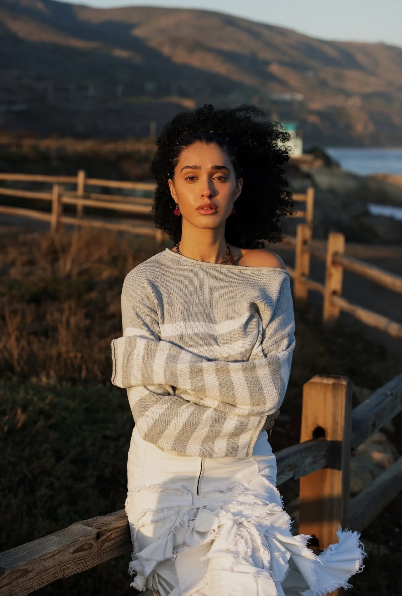 A woman with curly black hair and light brown skin standing outdoors during sunset, wearing a gray and white striped sweater and white distressed pants, with a coastal landscape in the background.