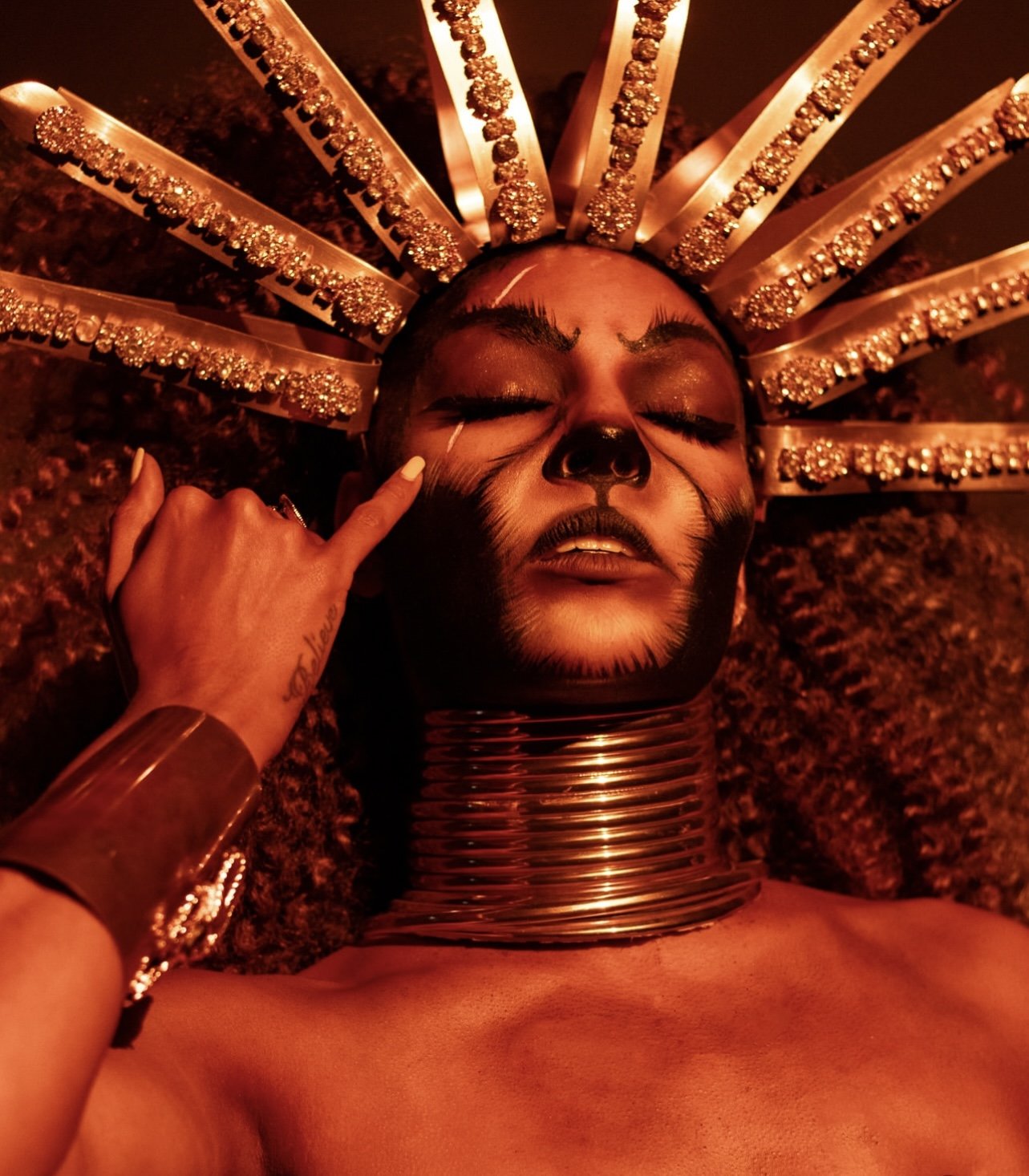 A person lying on the ground with their eyes closed, wearing an elaborate headpiece with gold and rhinestone decorations, and large metallic rings around the neck. The face is painted with makeup resembling a skeleton or Day of the Dead style, with a