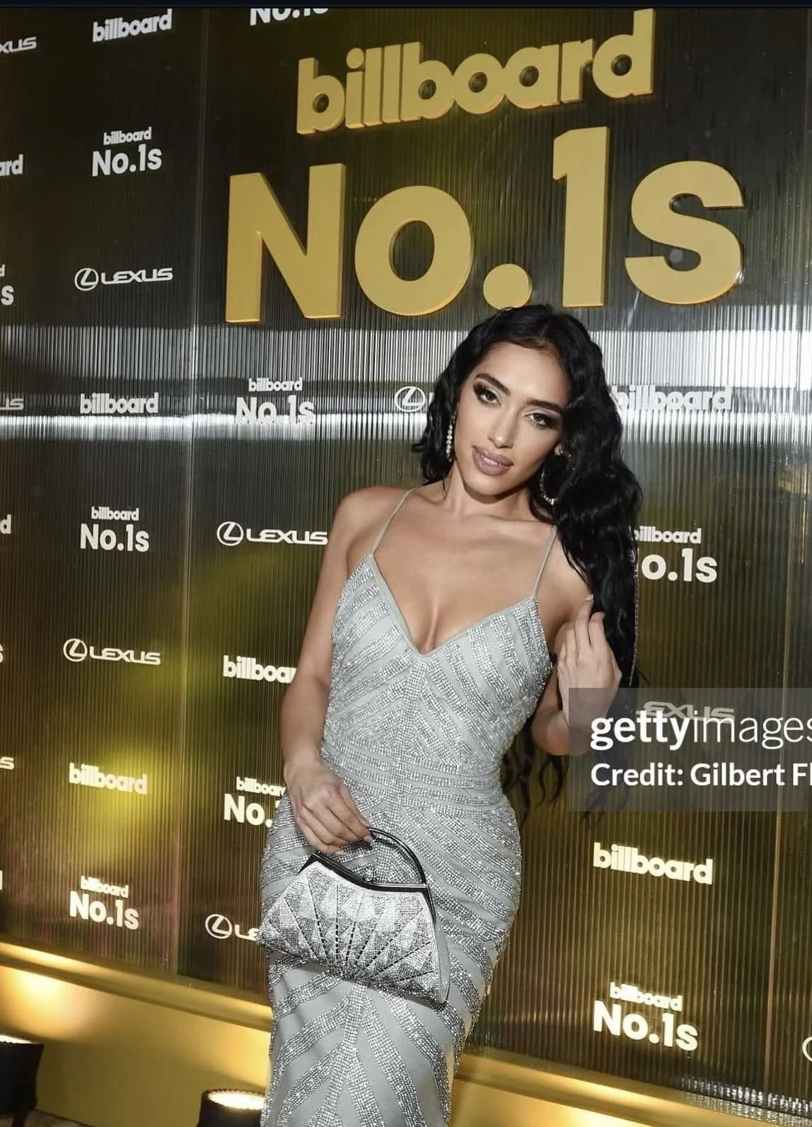 A woman in a silver, sparkling dress holding a small matching silver clutch posing at an event with a backdrop featuring the words 'billboard No.1s' and sponsor logos.