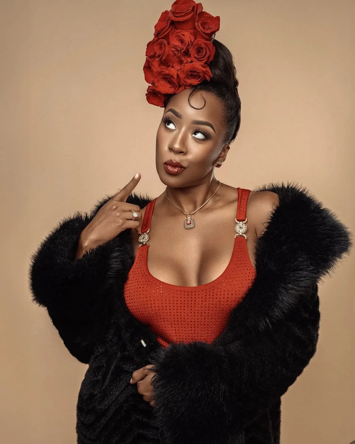 A woman with a tall headpiece made of red roses, wearing a red mesh top with a black fur coat, pointing to her head with her finger, against a neutral background.