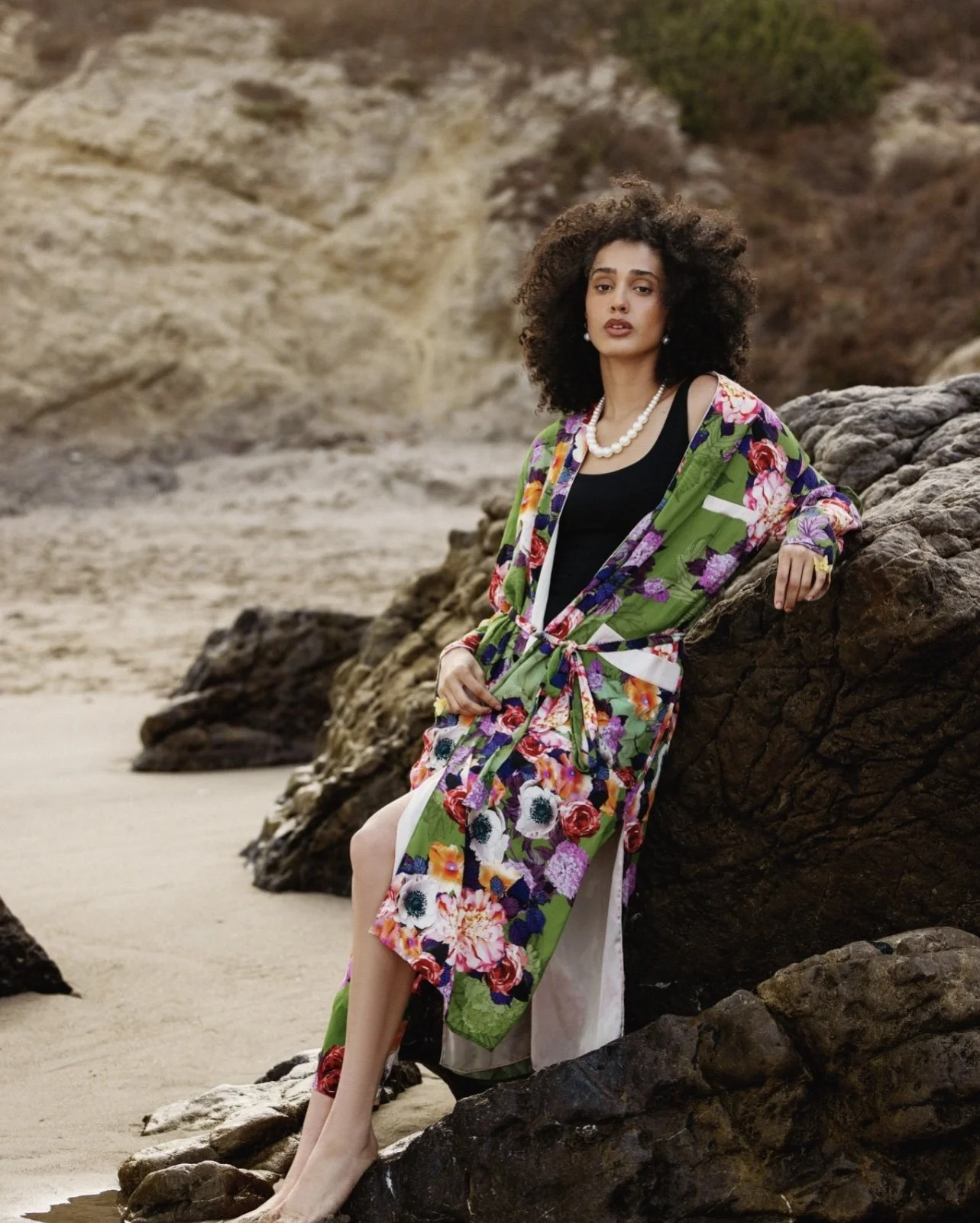 A woman with curly hair leaning on a large rock on a beach, wearing a black top, a floral jacket, a pearl necklace, and a ring.