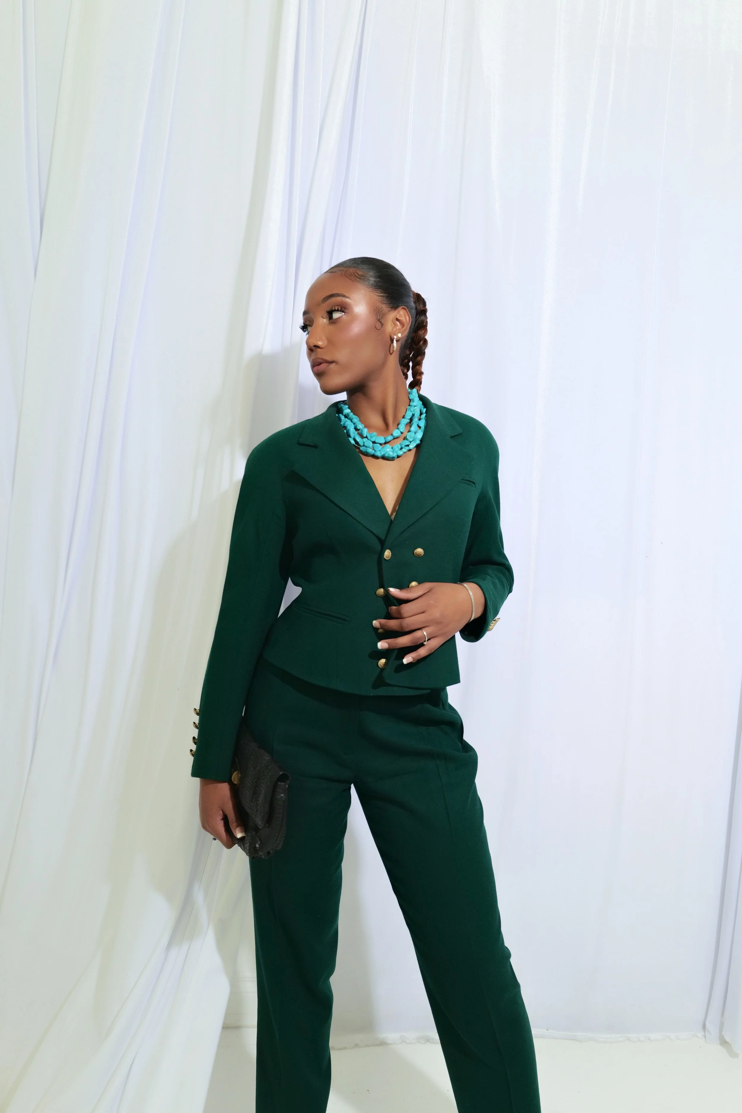 Woman in a green suit with gold buttons and a turquoise necklace, holding a black clutch, standing against a white curtain background.