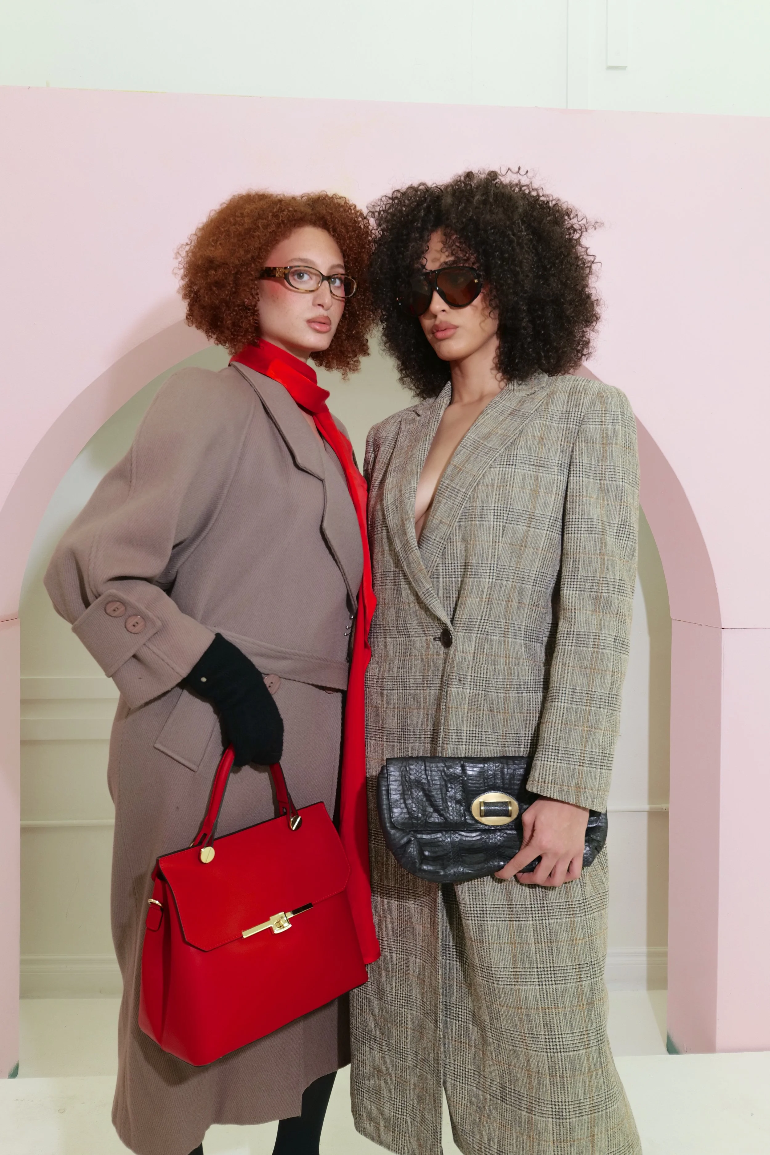 Two women posing against a pink arch background, wearing stylish suits and holding handbags.