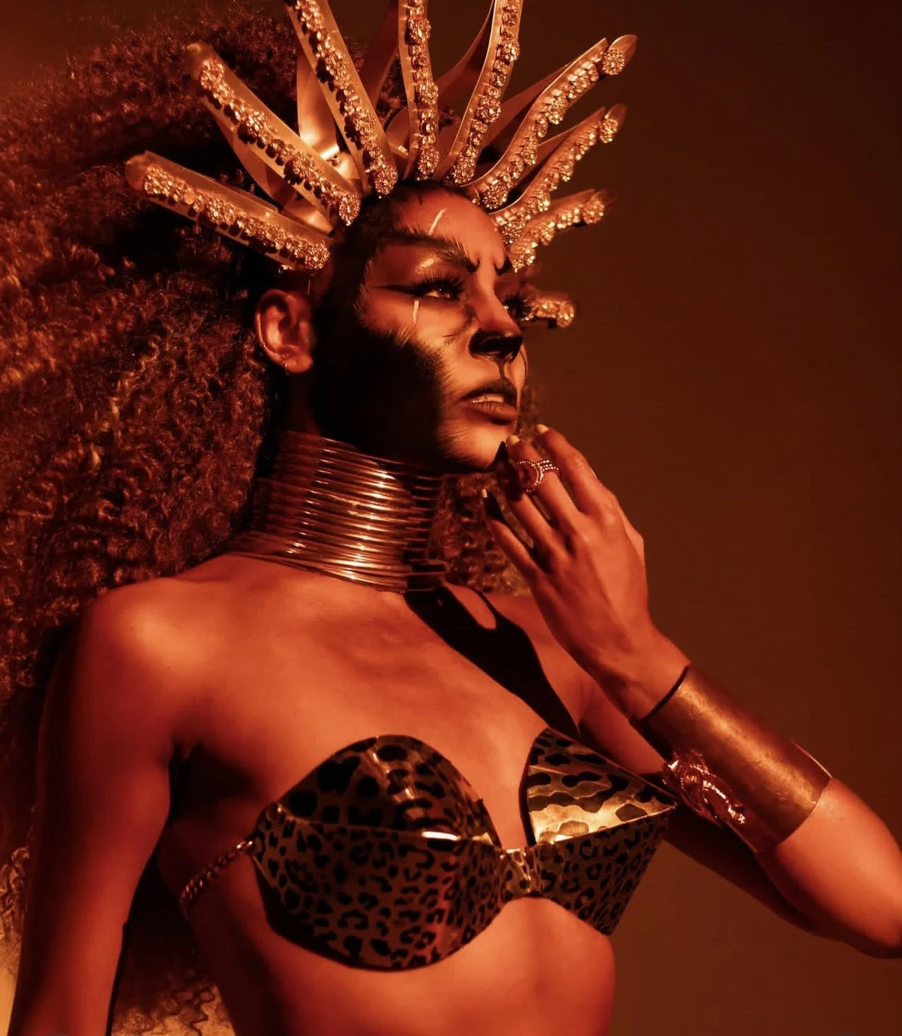 A woman with elaborate gold headpiece, face makeup, and metallic accessories, wearing a leopard print bra, standing against a warm, dimly lit background.