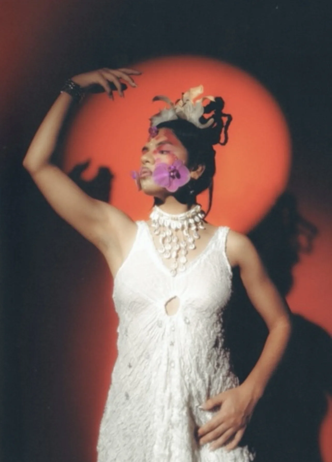 A woman in a white dress with a keyhole cutout, wearing pearls and a statement necklace, has flowers in her hair and a large purple flower covering her mouth, standing against a red and black background with a circular red light.