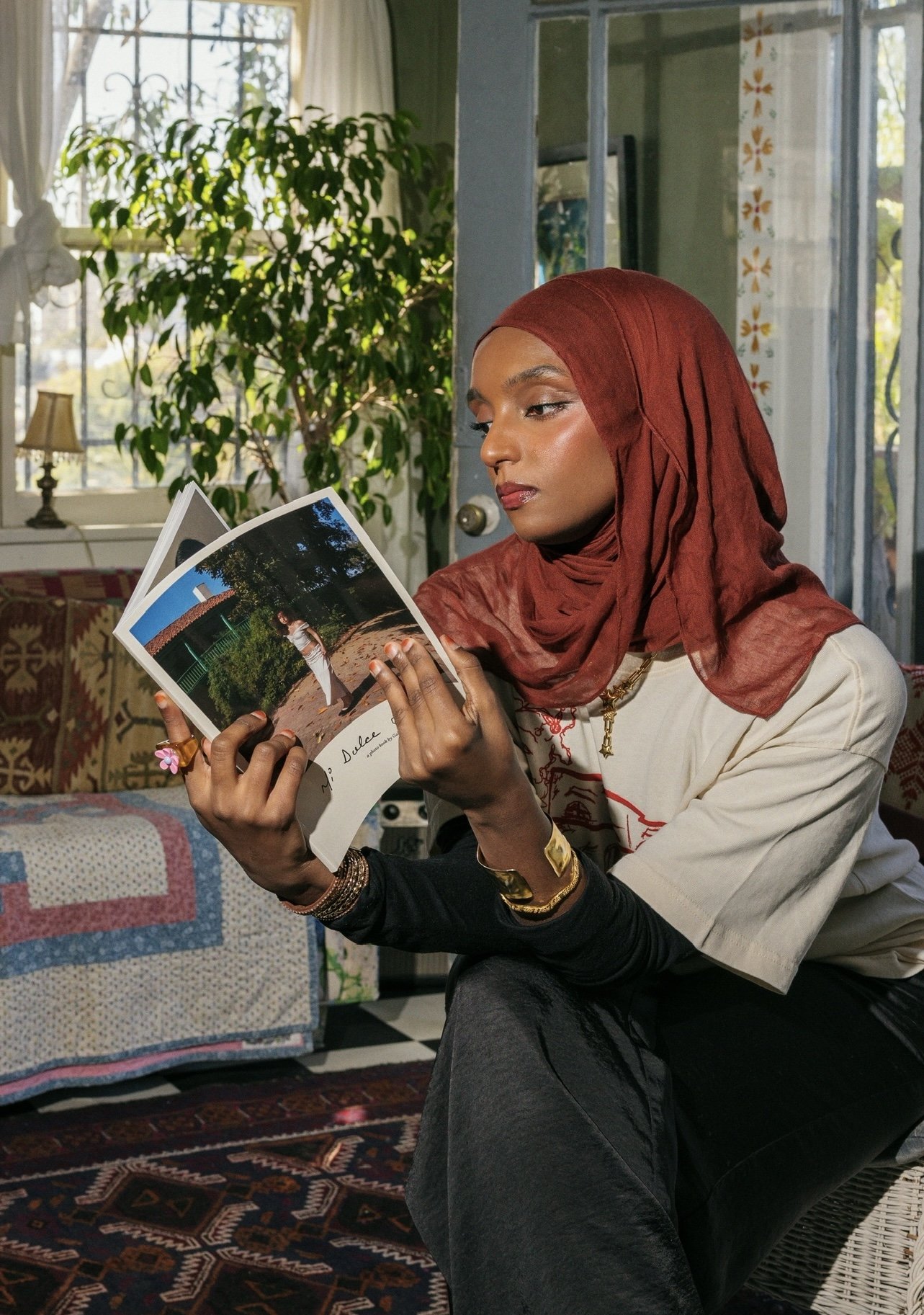 A woman wearing a red headscarf and beige shirt is sitting indoors, looking at a photo album. She has a thoughtful expression, with her lips slightly pursed. The background features a window with sunlight, a large green plant, and a cozy living room 