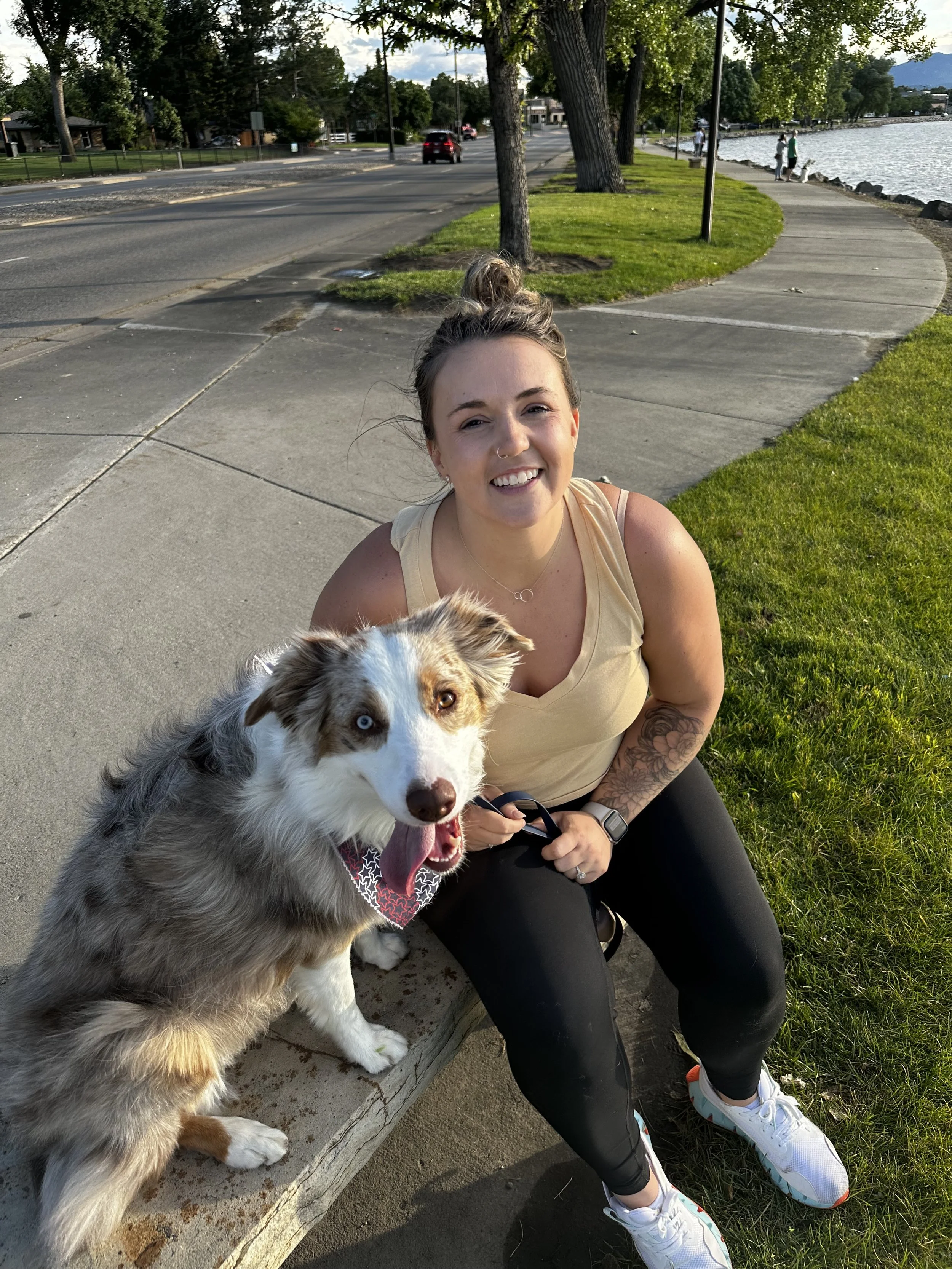 A smiling woman with a tattoo on her left arm sitting on a park bench with a dog that has heterochromatic eyes, one blue and one brown, near a body of water.