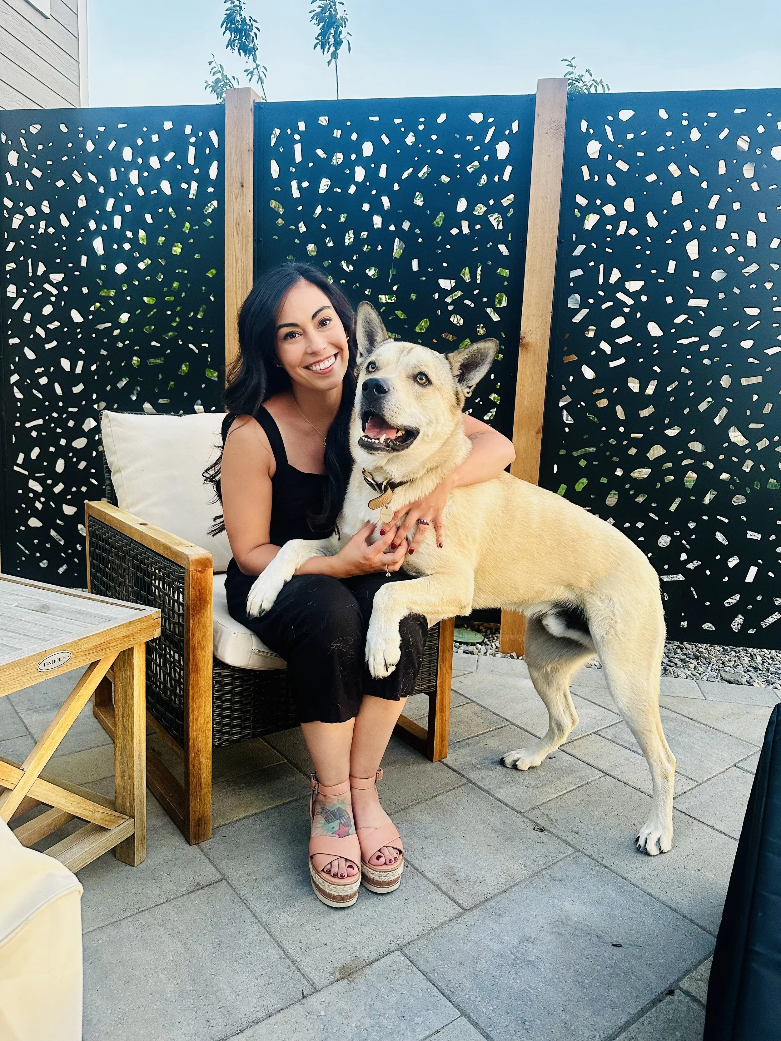 A woman sitting on a chair outdoors, smiling and hugging a large mixed-breed dog, with a black decorative privacy screen behind them.