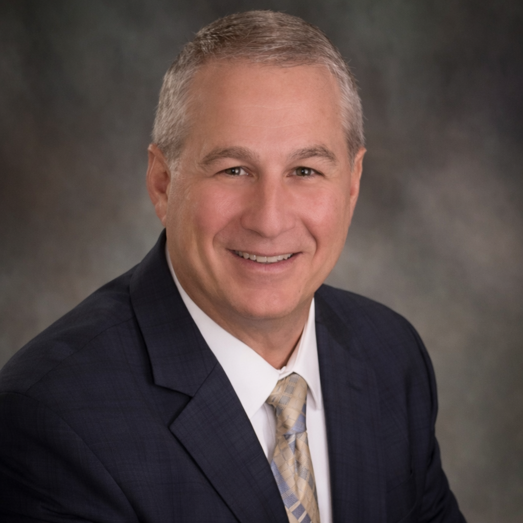 A middle-aged man in a dark plaid suit, white shirt, and light patterned tie, smiling in a professional studio portrait.