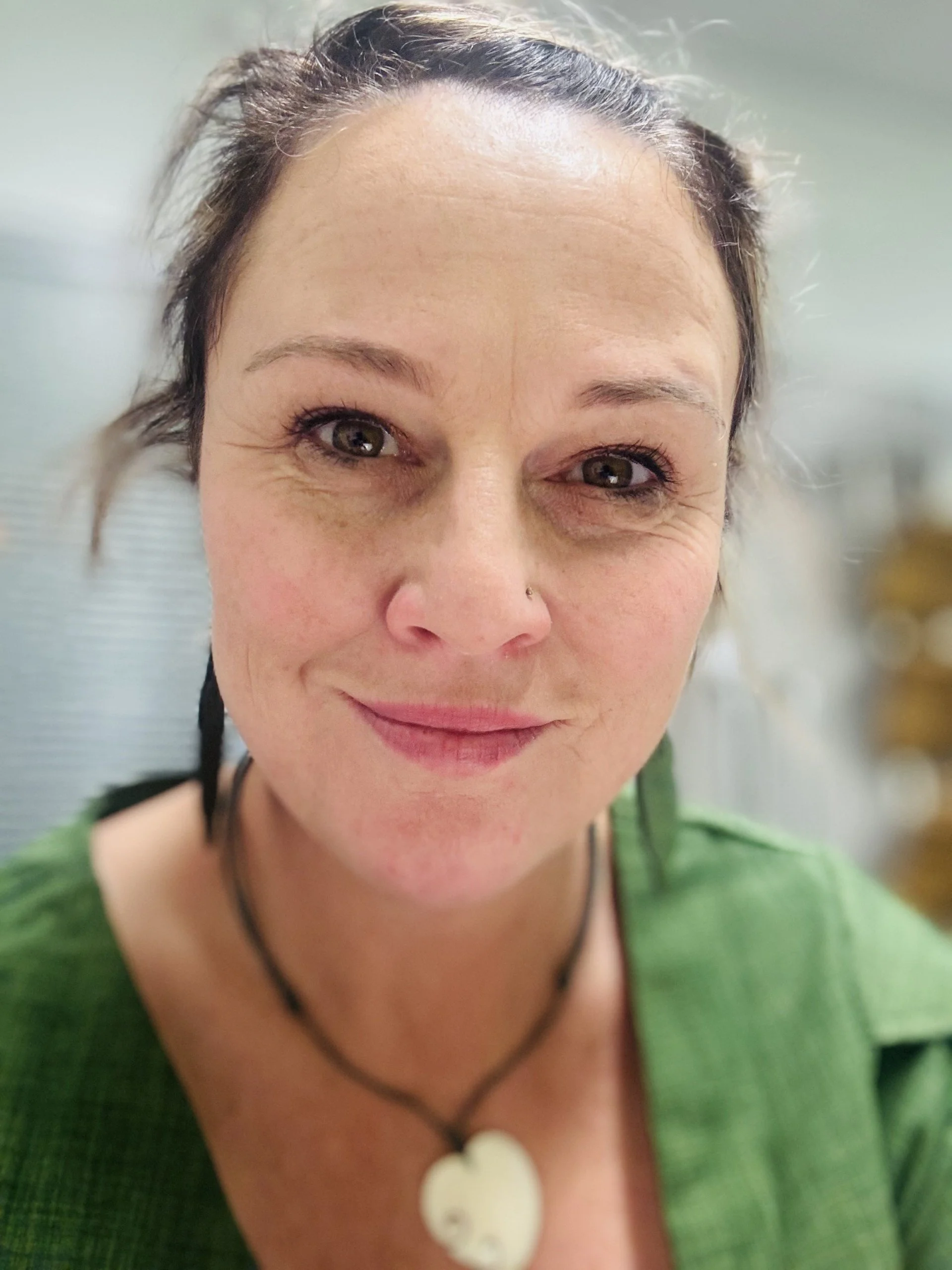 Close-up of a smiling woman with dark hair and light skin, wearing a green top and a heart-shaped pendant necklace.