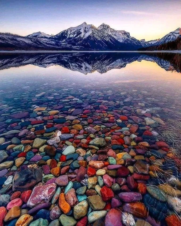 Colorful pebbles on the shore of a clear lake with snow-capped mountains reflected in the water during sunset.