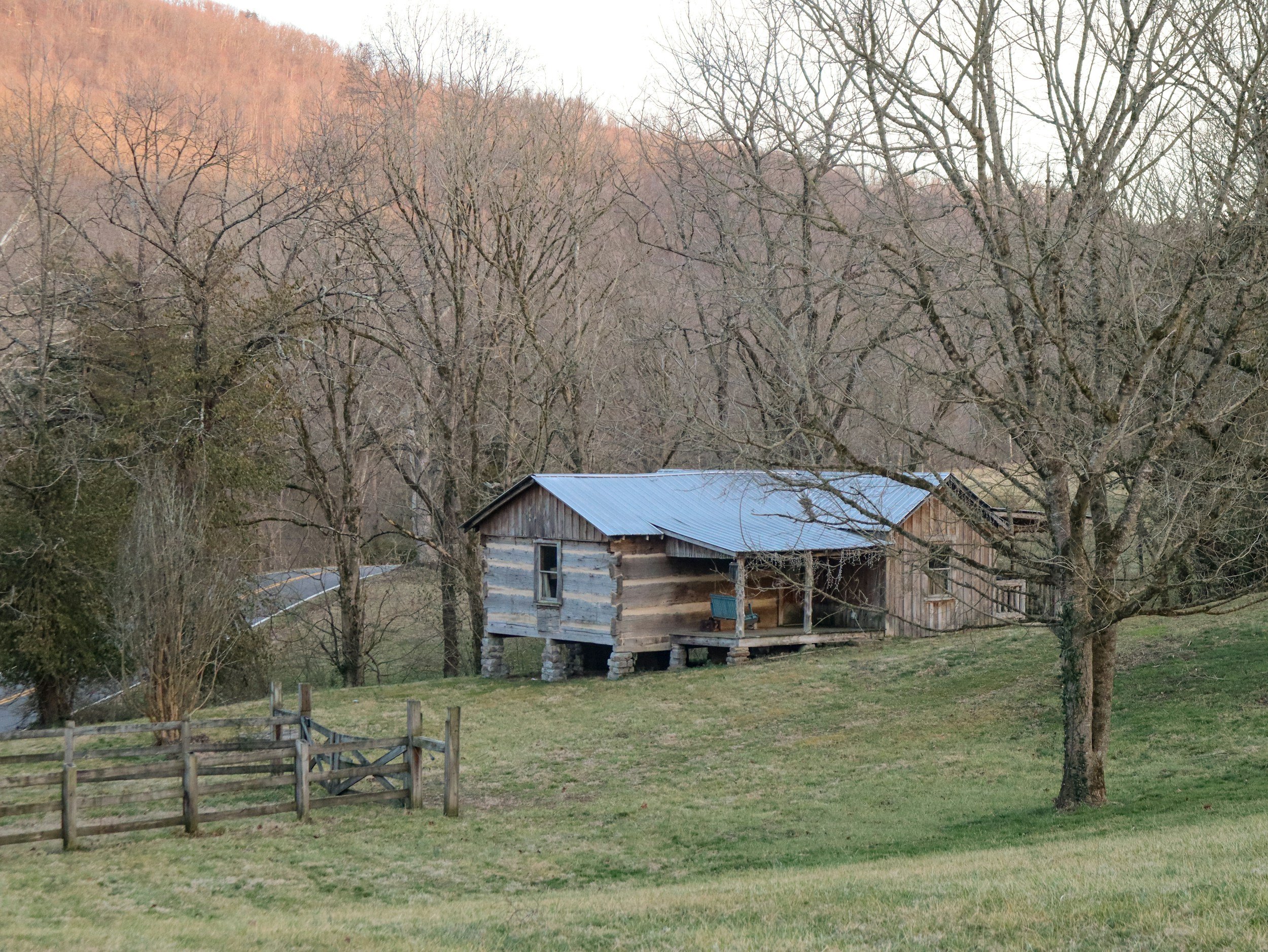 A rustic farm building with a metal roof situated on a grassy hillside, surrounded by leafless trees and a wooden fence, with hills in the background.