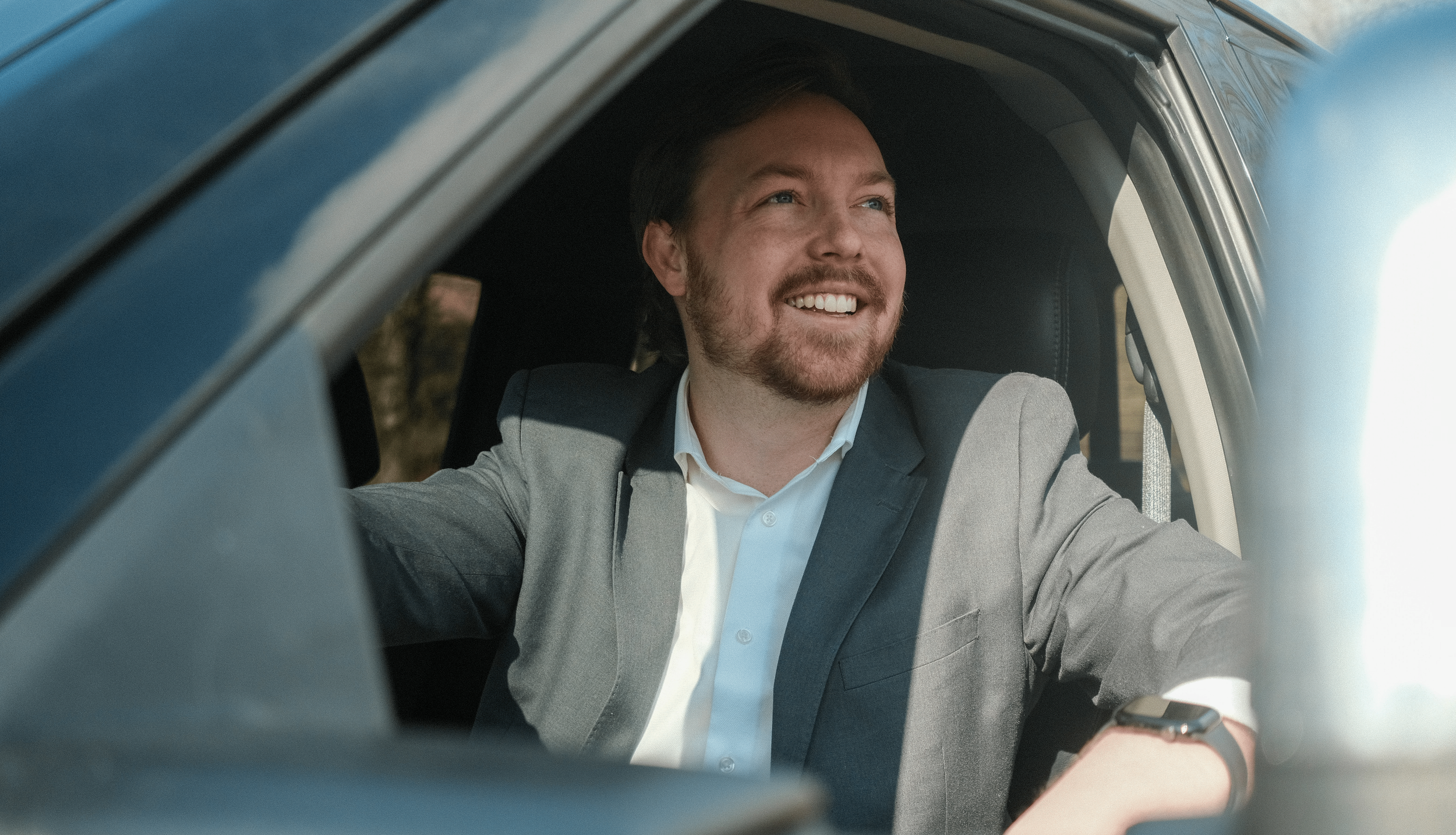 Man with beard smiling and looking out the car window, wearing a gray suit jacket and a white shirt, with a smartwatch on his wrist.
