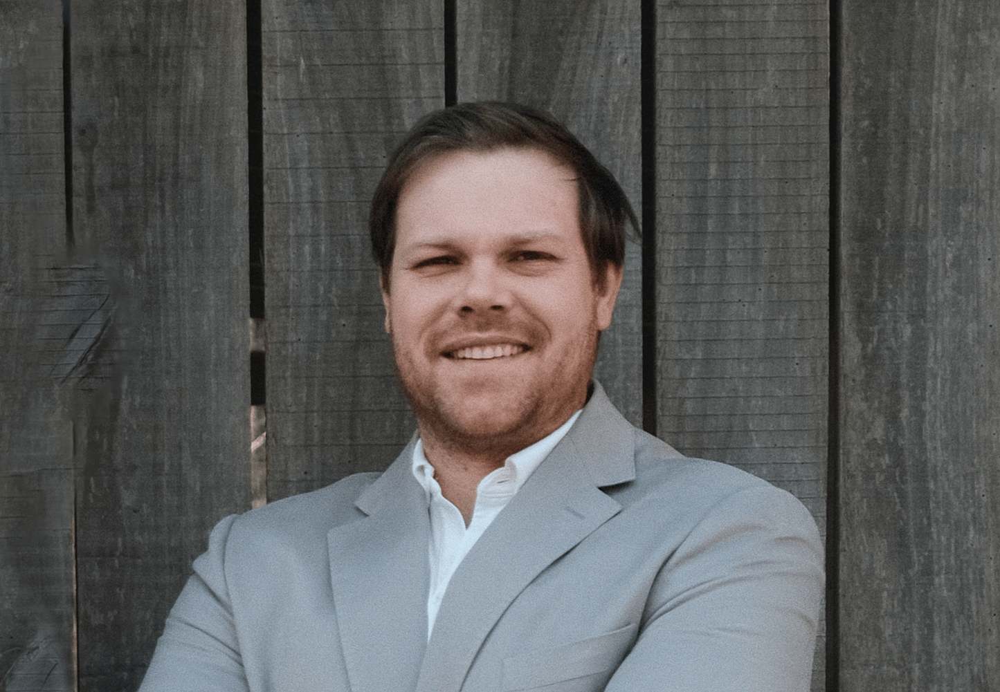 Portrait of a man smiling in a light gray suit in front of a wooden fence.
