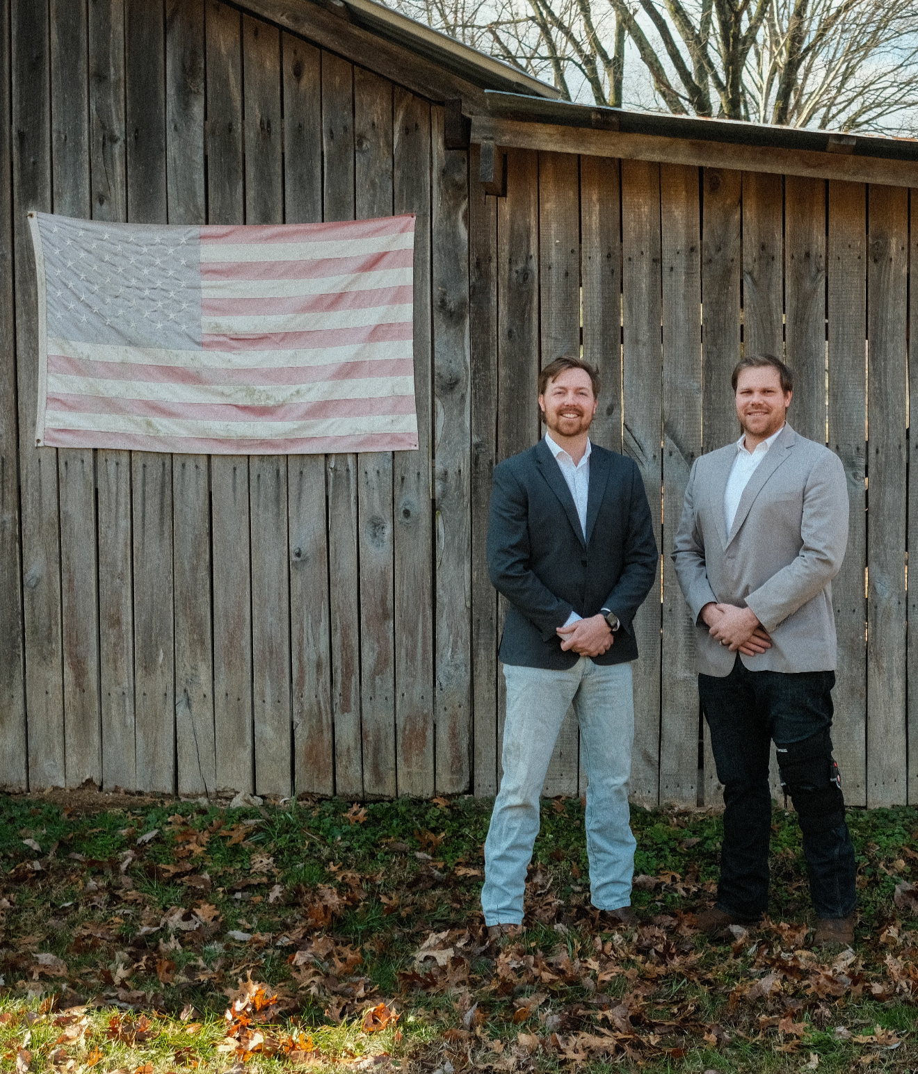 Two men in suits standing outdoors in front of a wooden fence with an American flag hanging on it.