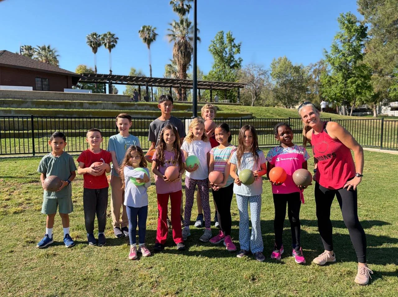 Group of children and a coach in a park, holding various sports balls on a sunny day.