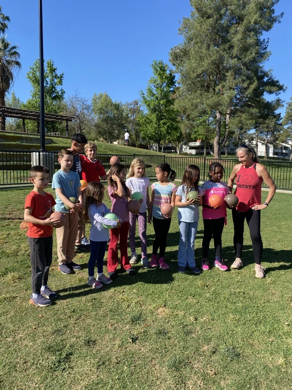 Group of children and woman outdoors holding basketballs, in a park on a sunny day.