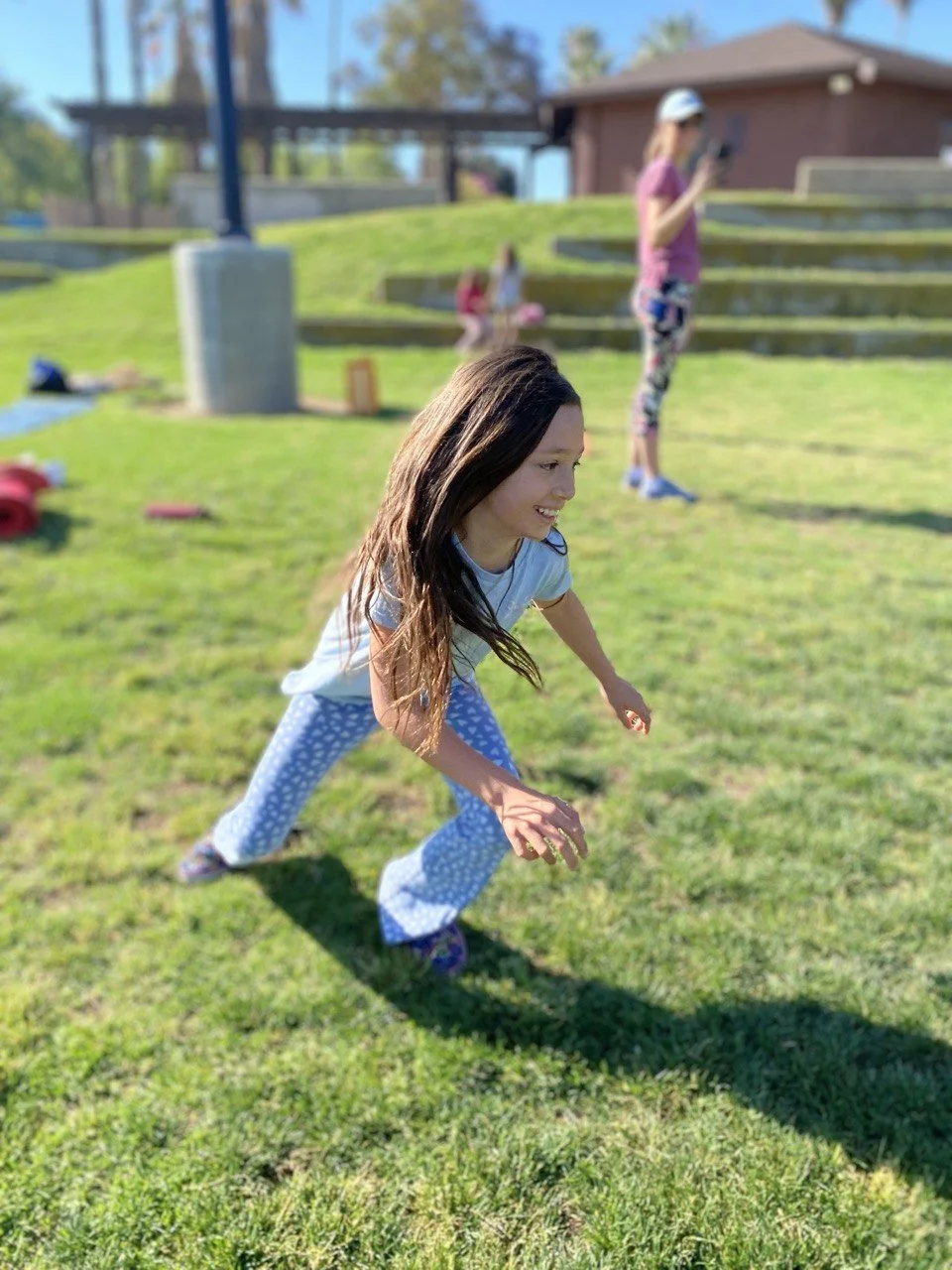 A young girl in blue polka dot pajamas crouches in a yard with grass, smiling, as she plays outdoors on a sunny day. In the background, there are two other girls, one standing and looking at her phone, and the other sitting on the steps of a house.