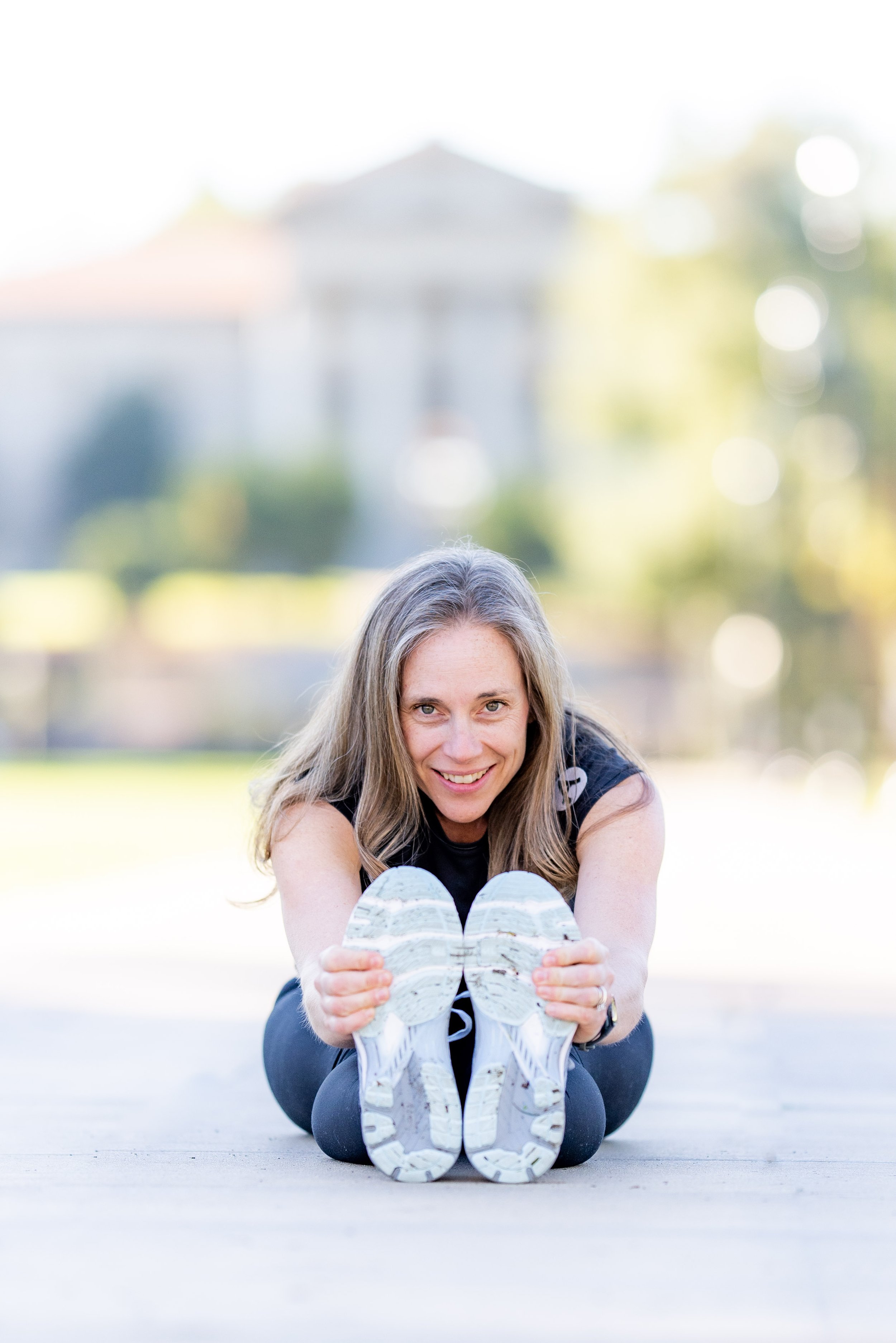 Woman with long blonde hair smiling, wearing black athletic wear, sitting on the ground and holding her white running shoes facing the camera, with a blurred outdoor background and greenery.