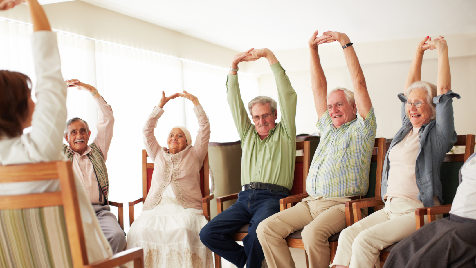A group of seniors participating in a seated stretching or yoga class, raising their arms above their heads and smiling in a bright, well-lit room.