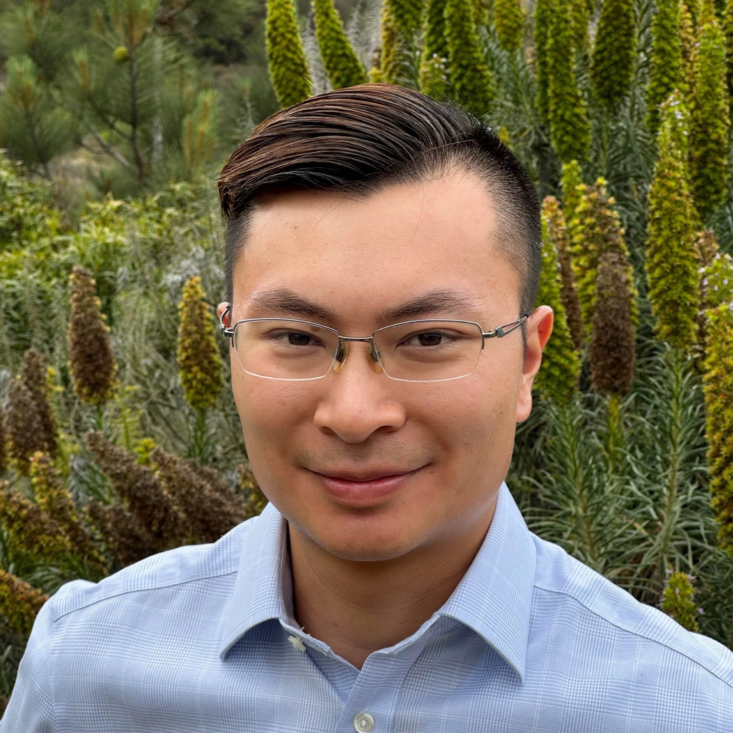 A person with short, styled hair and glasses, dressed in a light blue shirt, standing outdoors in front of green plants and yellow flowers.