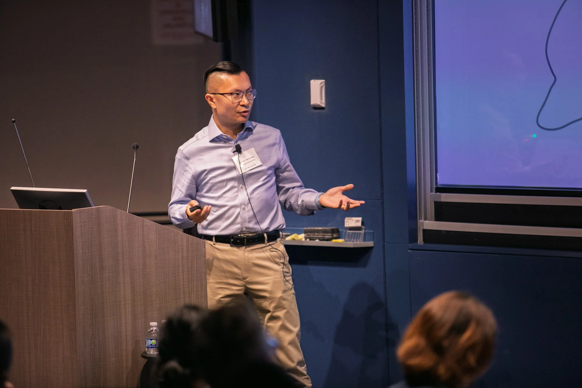 A man with glasses and a short haircut giving a presentation in a conference room, standing at a podium with a microphone and a large screen behind him displaying a diagram. Audience members are visible in the foreground.