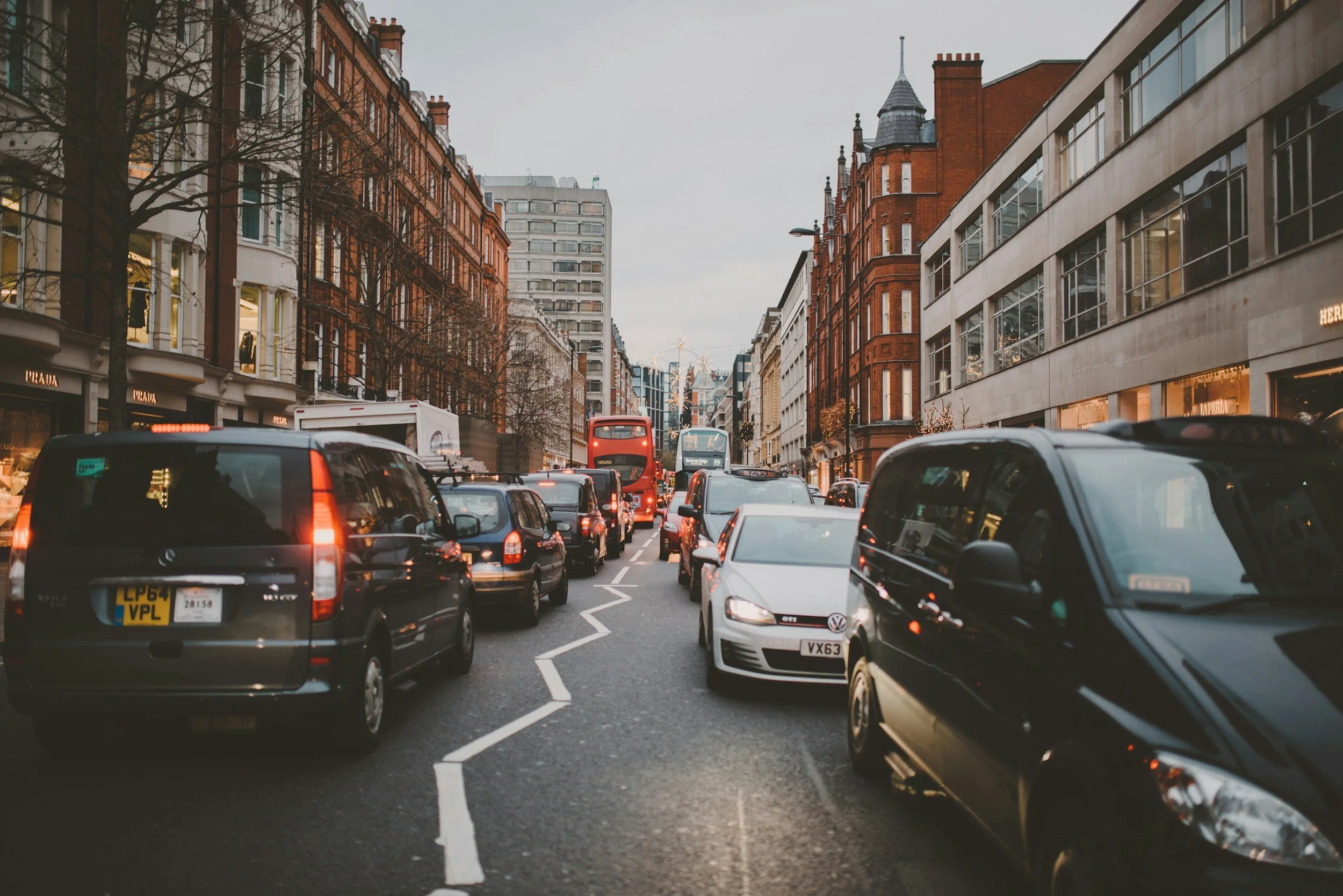 Traffic congestion on a city street with cars, a red double-decker bus, and buildings with shops and windows in the background.