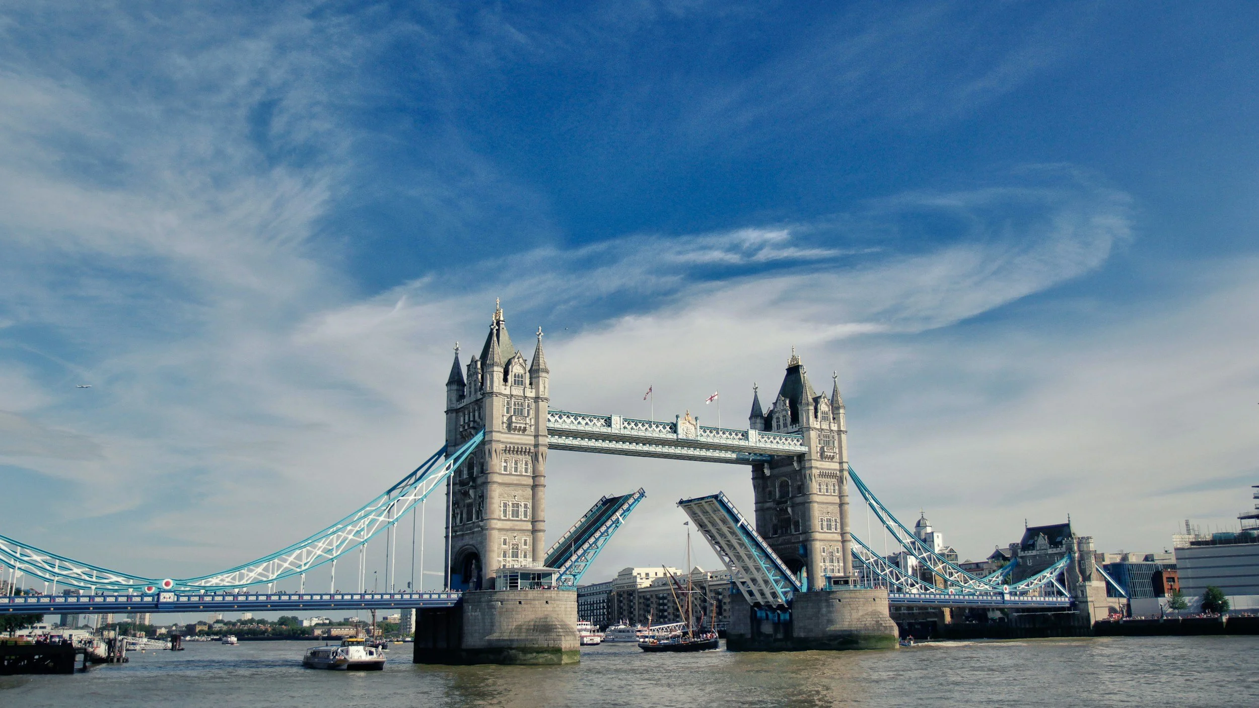 The Tower Bridge in London with a clear blue sky and some clouds, boats on the River Thames, and some buildings in the background.