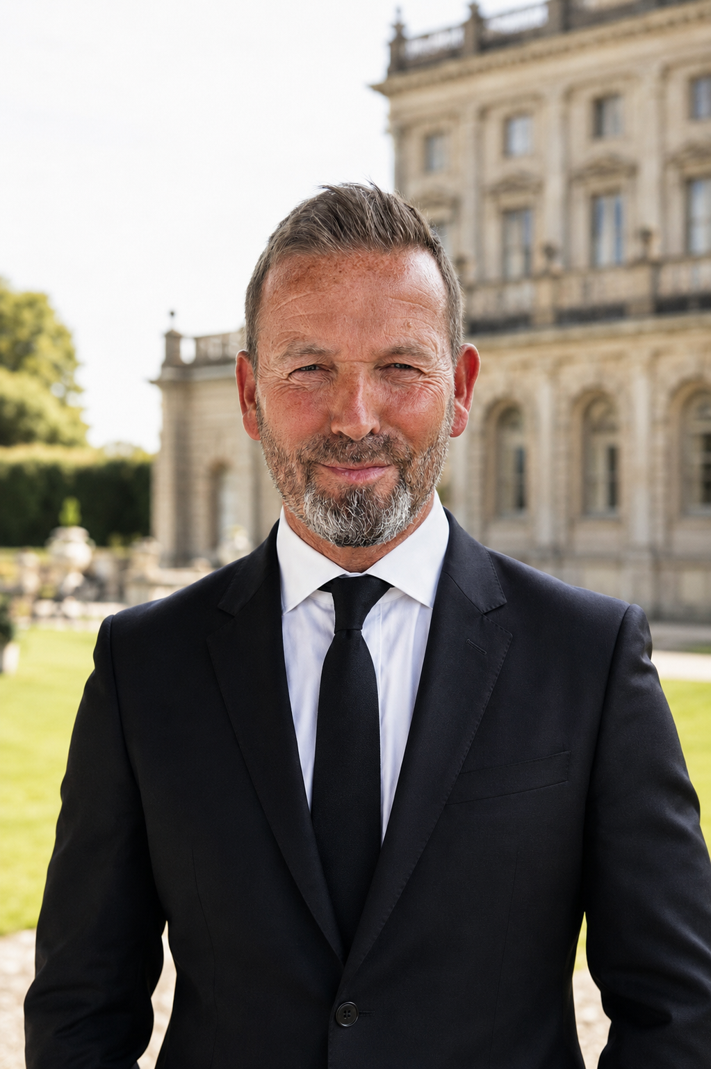A middle-aged man with a beard in a black suit and tie standing outside in front of a historic building with columns and ornate architecture, smiling.