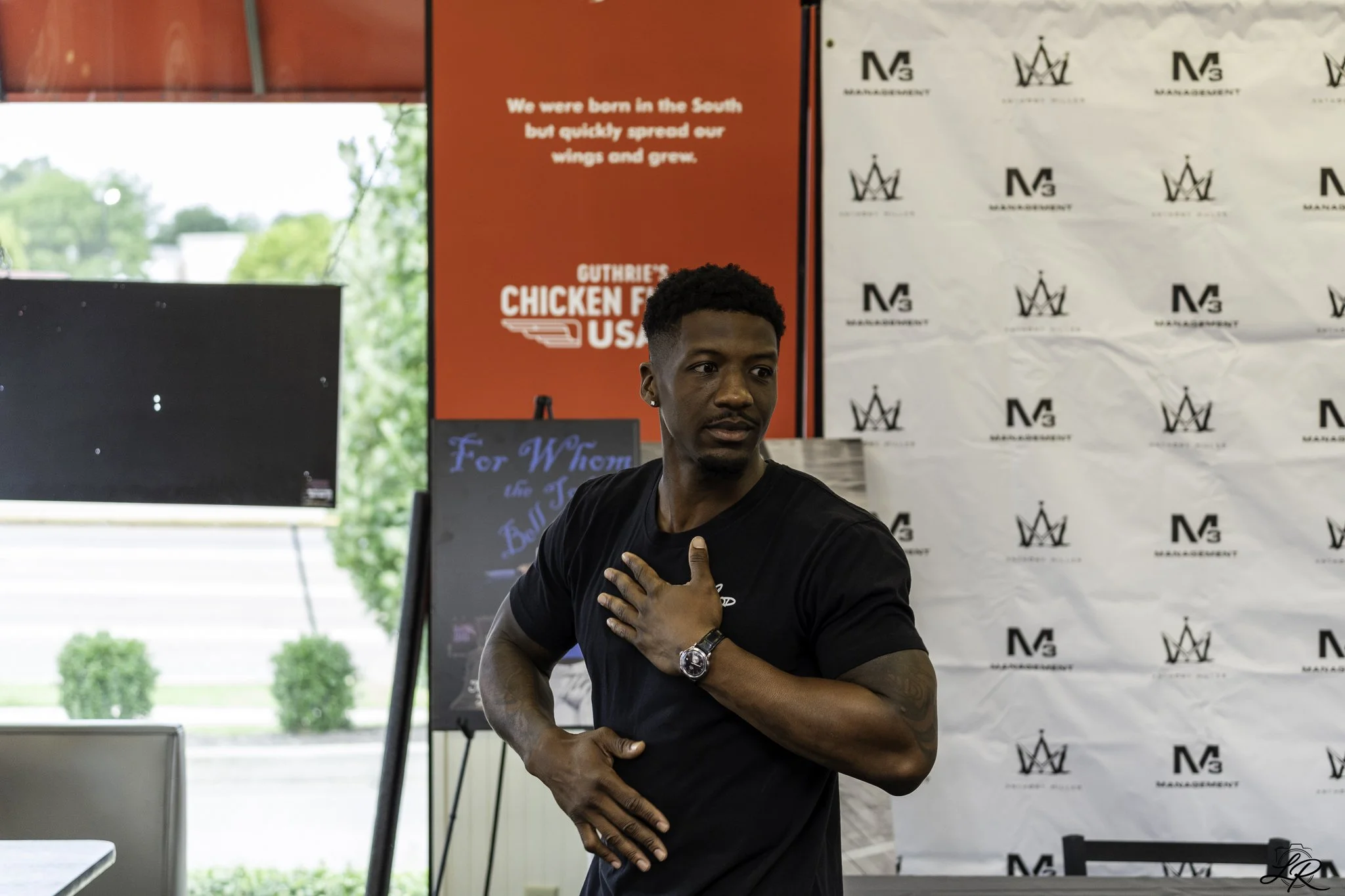 A young man in a black t-shirt with a watch on his left wrist is standing indoors. He has a hand on his chest and appears to be singing or speaking passionately. Behind him, there are banners with logos and text, including a red sign that says 'We we