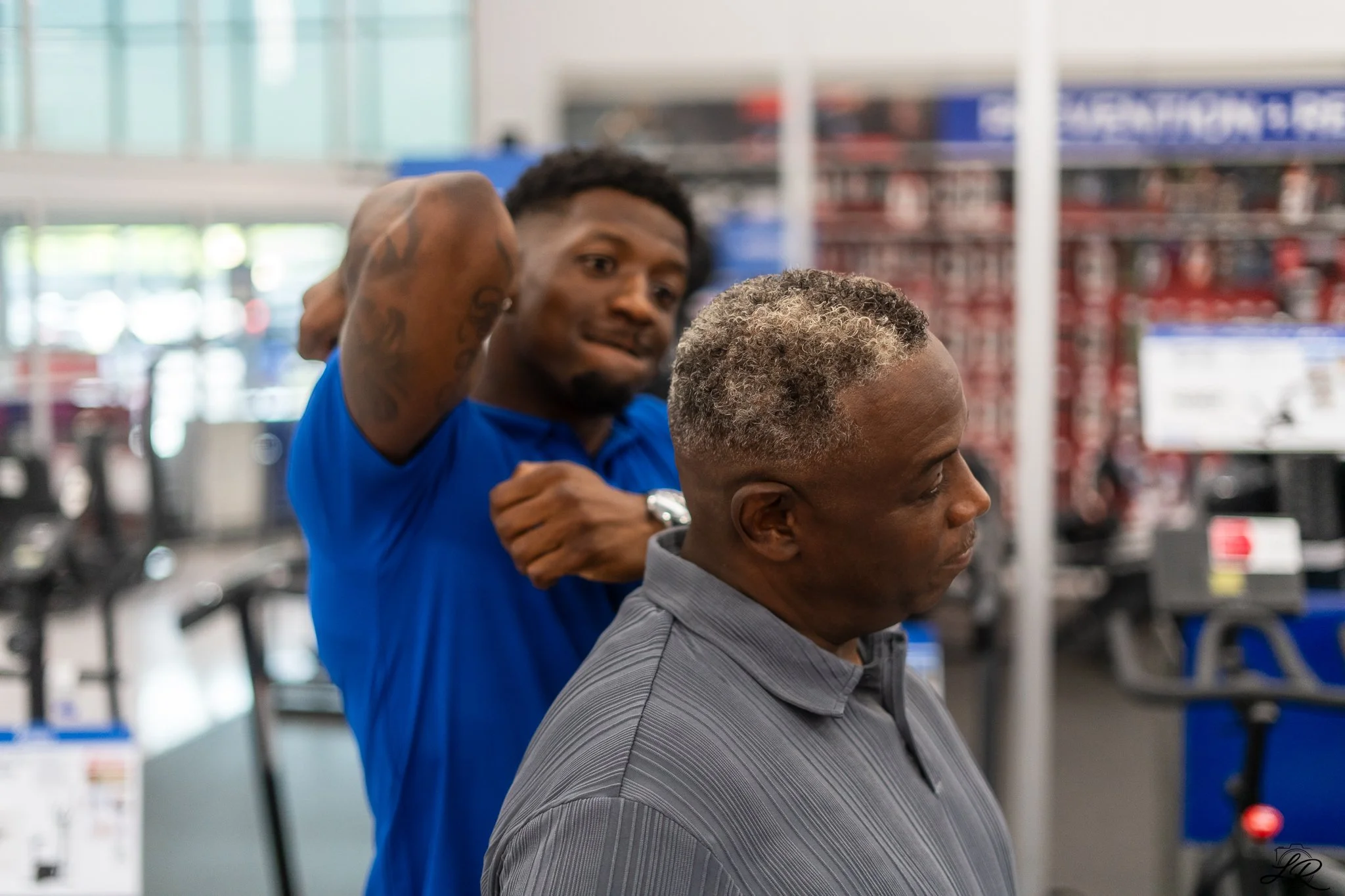 A man with short curly gray hair sits in a salon chair getting a haircut from a young barber with dark curly hair and tattoos on his arm, inside a store with shelves and banners in the background.