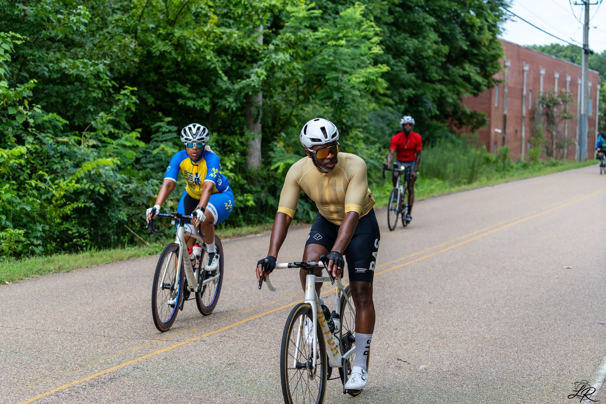 Three cyclists riding on a paved road surrounded by green trees and bushes, with a brick building and utility poles in the background.