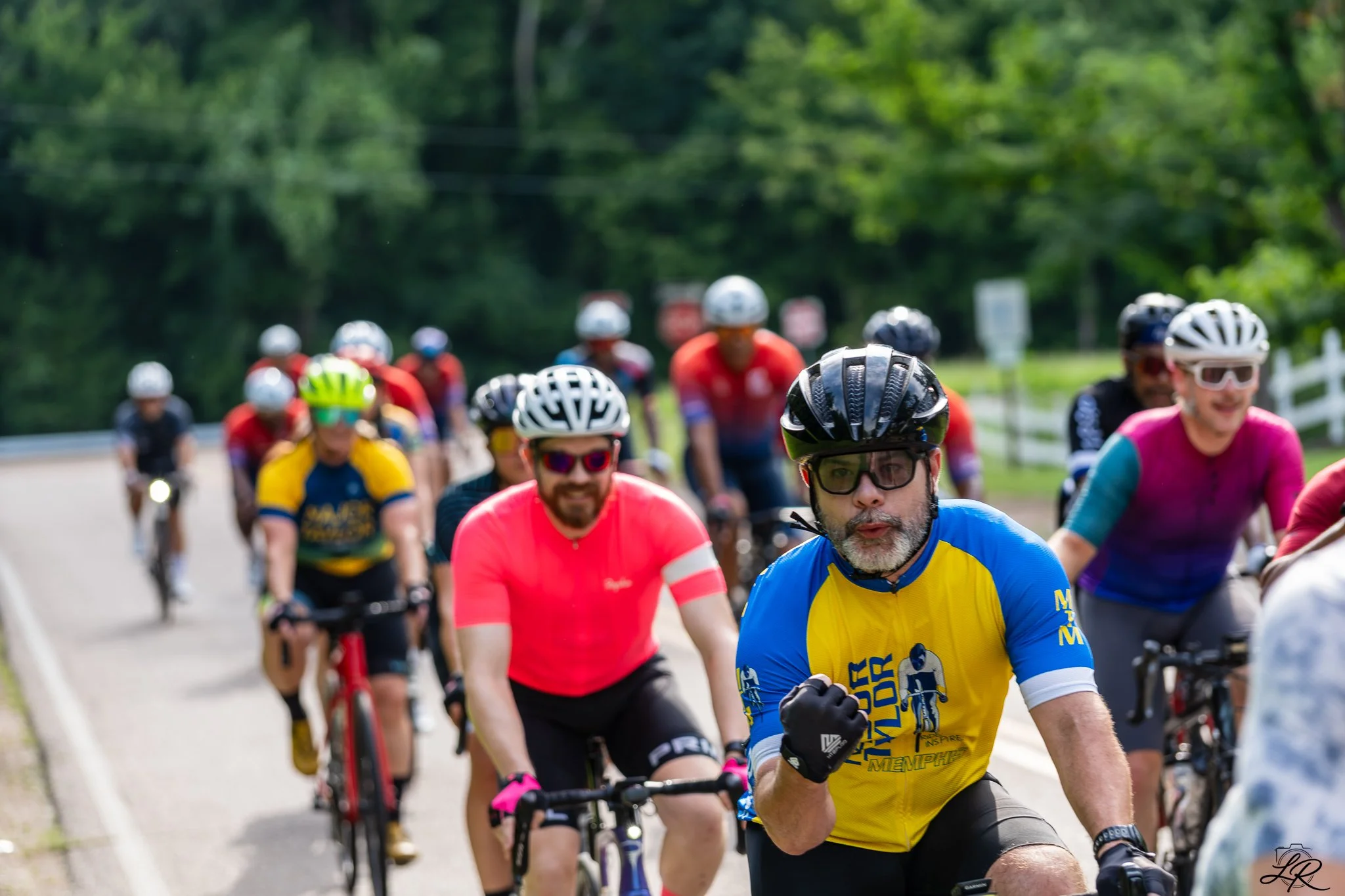 Group of cyclists riding on a rural road surrounded by green trees, some wearing colorful jerseys and helmets.
