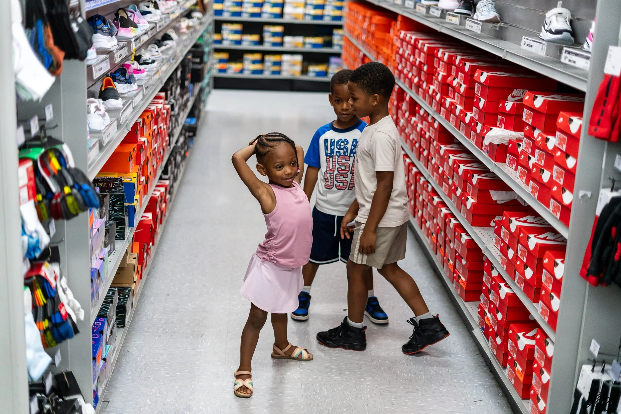 Three children standing in a shoe aisle of a store, looking at sneakers. The girl in a pink dress is scratching her head, while the two boys are looking at her. The shelves are filled with Nike shoes and other brands.