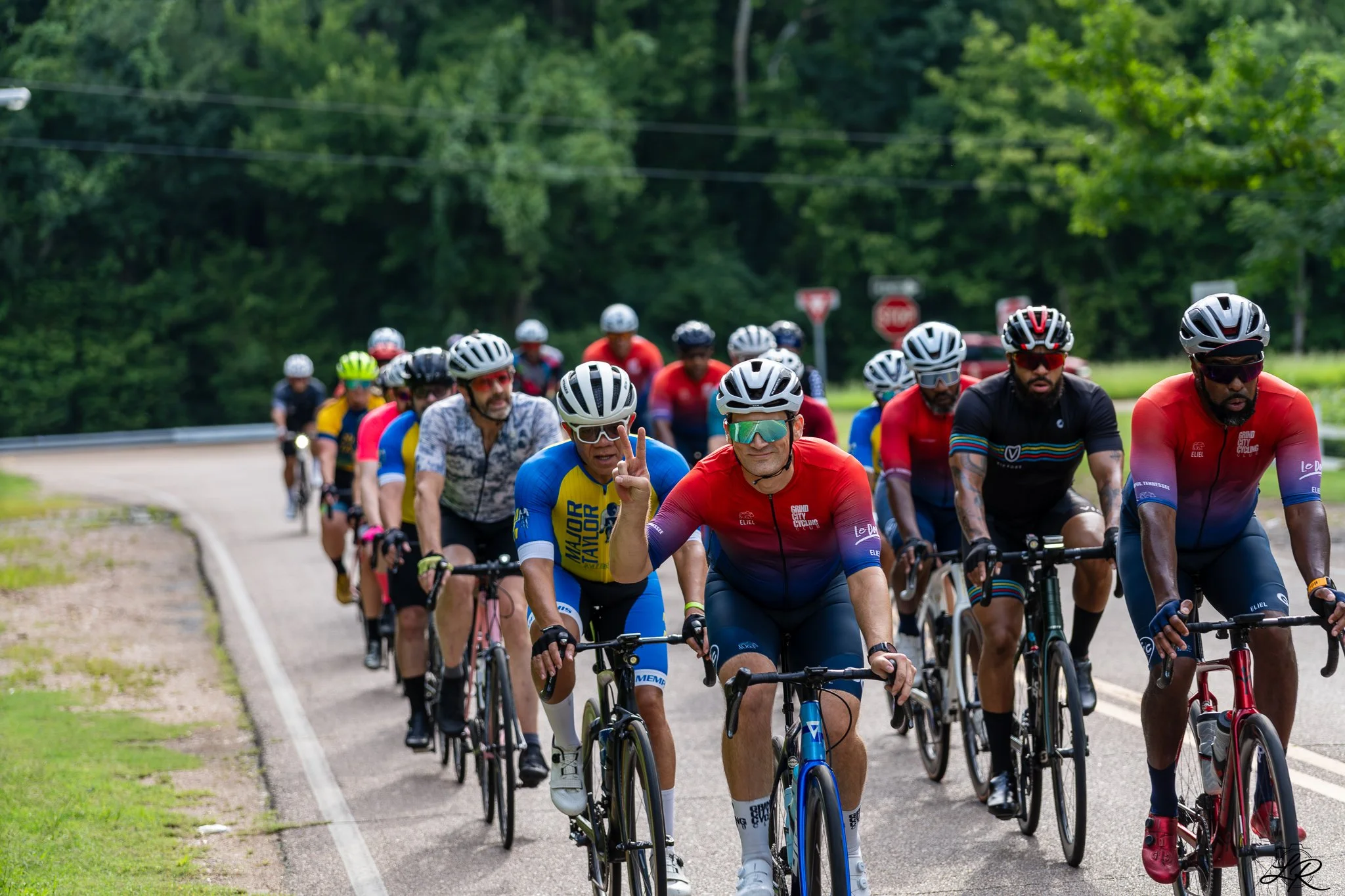 A group of cyclists riding on a road surrounded by trees, with some cyclists wearing helmets and sunglasses, one cyclist making a peace sign.