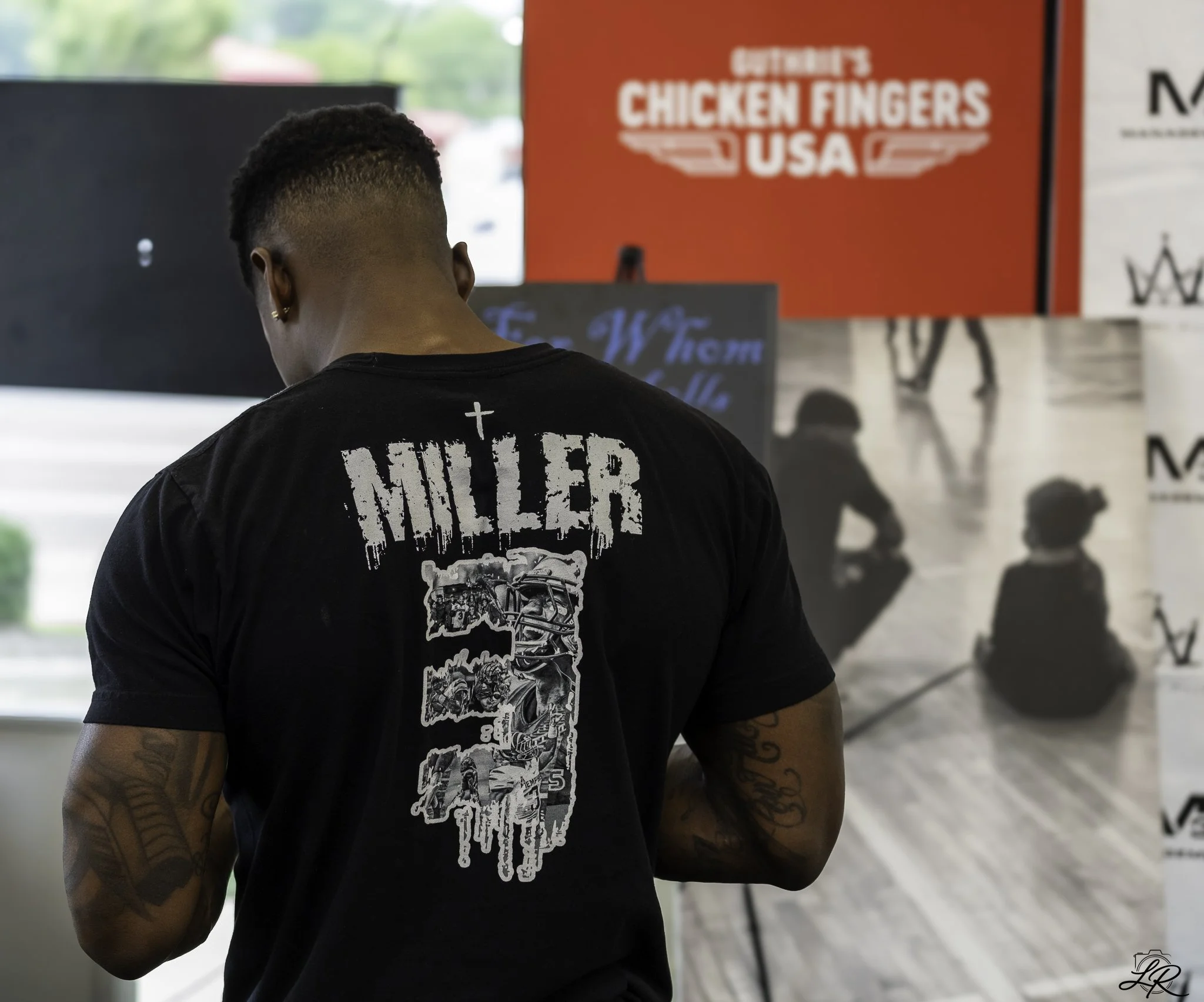 Back of a person with tattooed arms, wearing a black T-shirt with a graphic design and text, standing in front of a red sign that reads "Chicken Fingers USA". The person has short, curly hair and is wearing earrings.